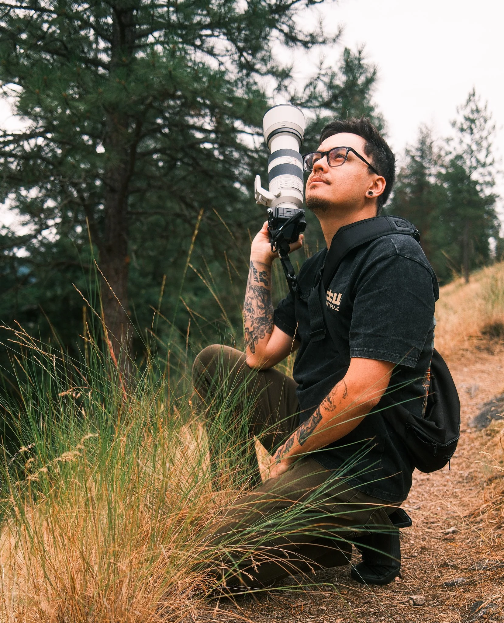 A man with tattoos and glasses sits outdoors in a forest, holding a large camera telephoto lens mounted on a camera body, looking into the distance.