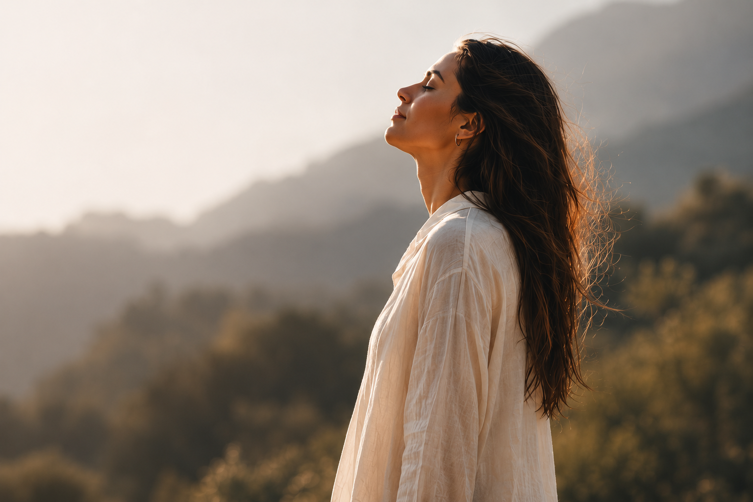 A woman with long dark hair and a light-colored shirt stands outdoors with her eyes closed and face tilted upward, enjoying the sunlight with mountains and trees in the background.