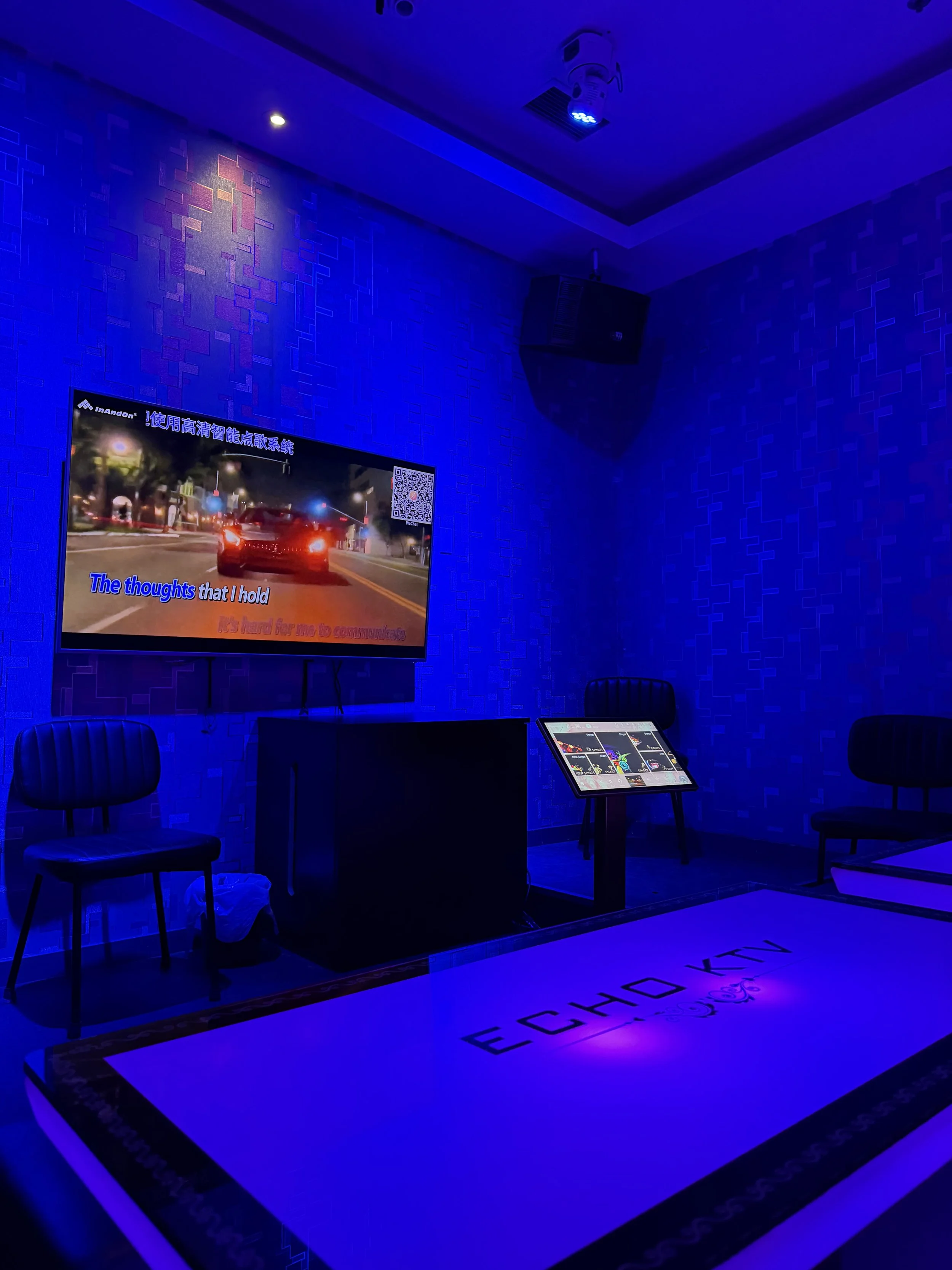 A dark room with blue lighting, containing a television displaying a car and street scene with karaoke lyrics, two chairs, a table with a tablet, and a large welcome mat on the floor.