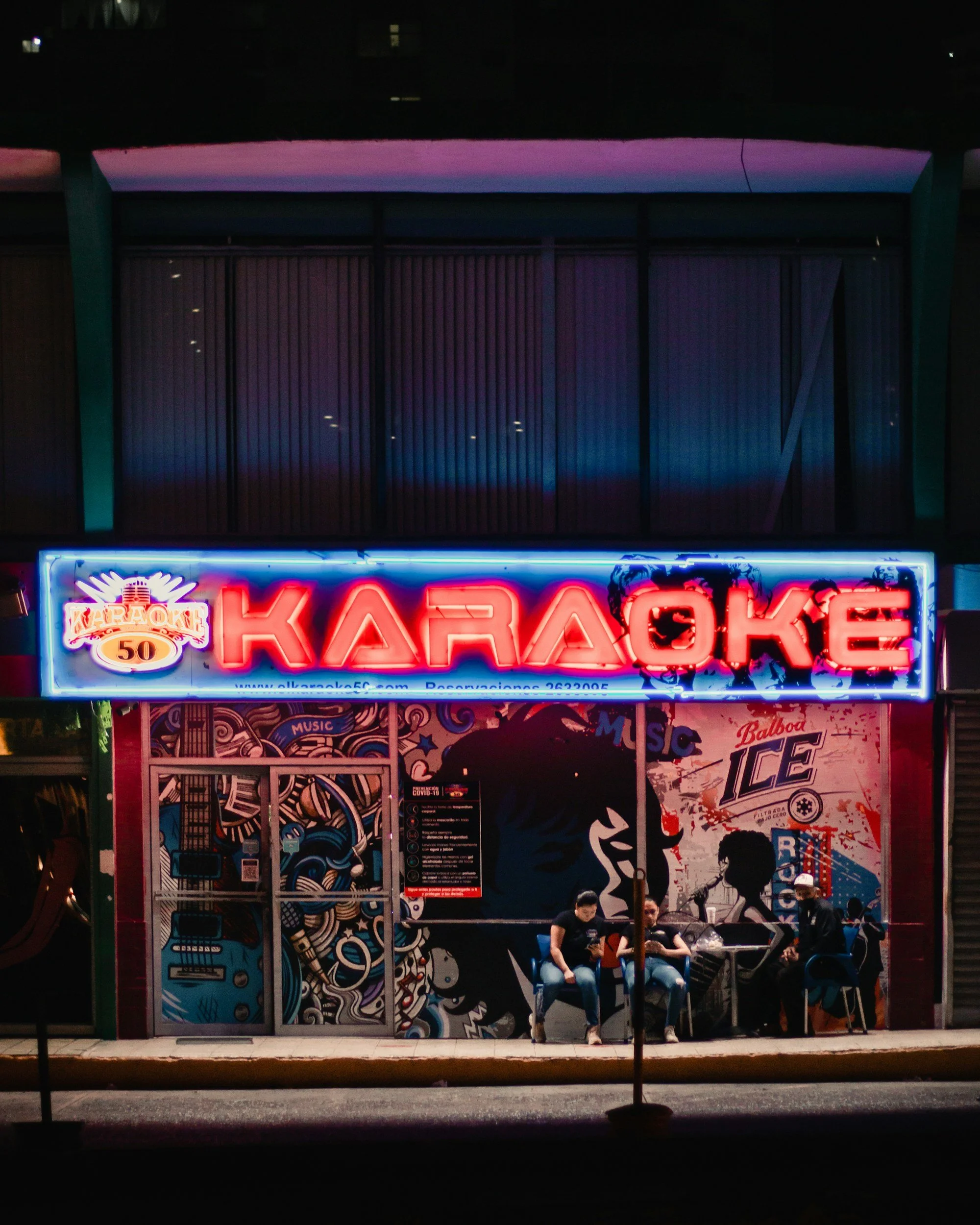 Neon sign advertising karaoke, featuring colorful graphics and silhouettes of musicians. Several people sit beneath the sign, illuminated by its glow.
