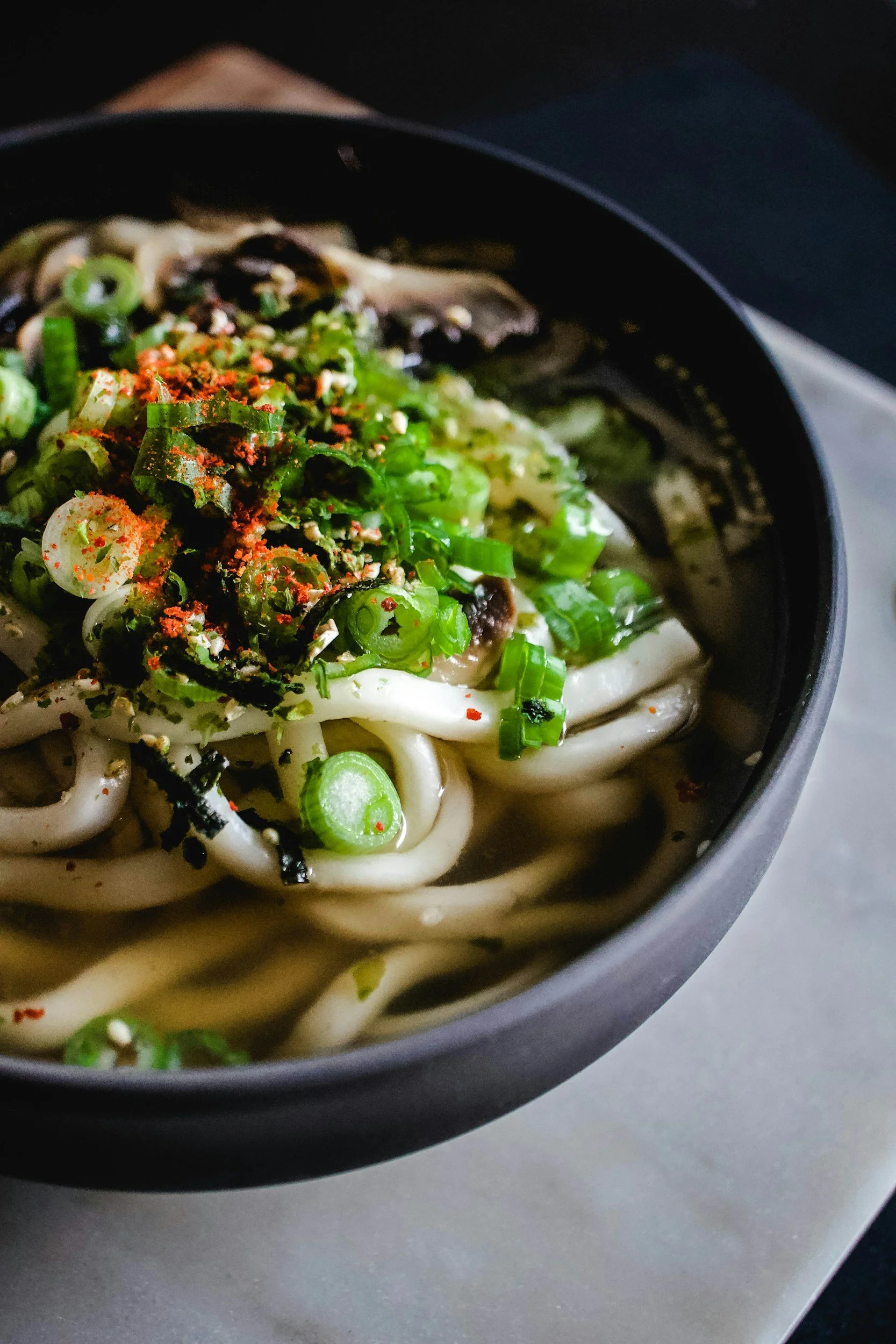 Close-up of a bowl of ramen noodles topped with chopped green onions, sliced mushrooms, red chili flakes, and sesame seeds.