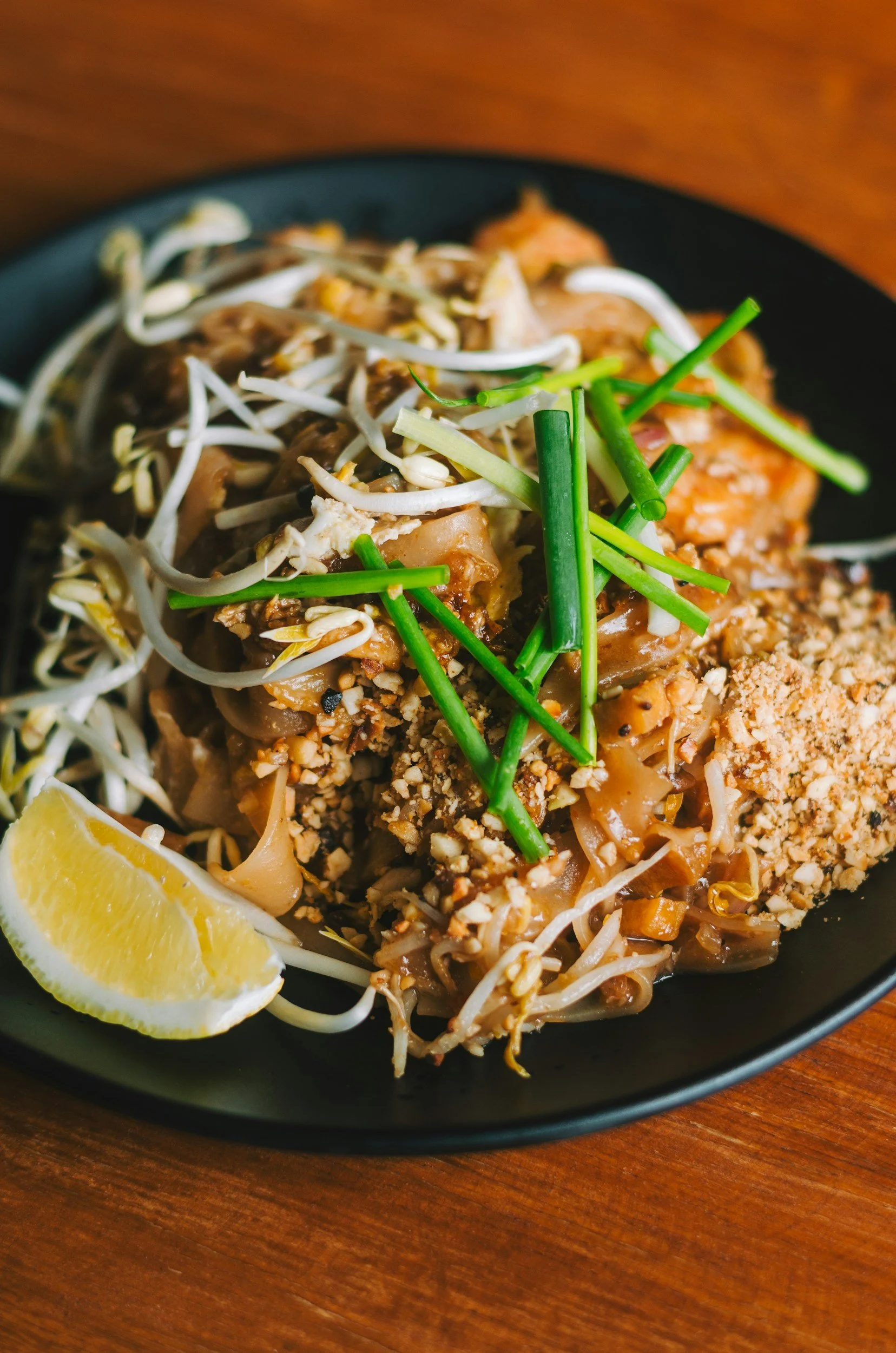 Close-up of a black bowl of Asian noodles topped with bean sprouts, chopped peanuts, chopped green onions, and a lemon wedge on a wooden table.