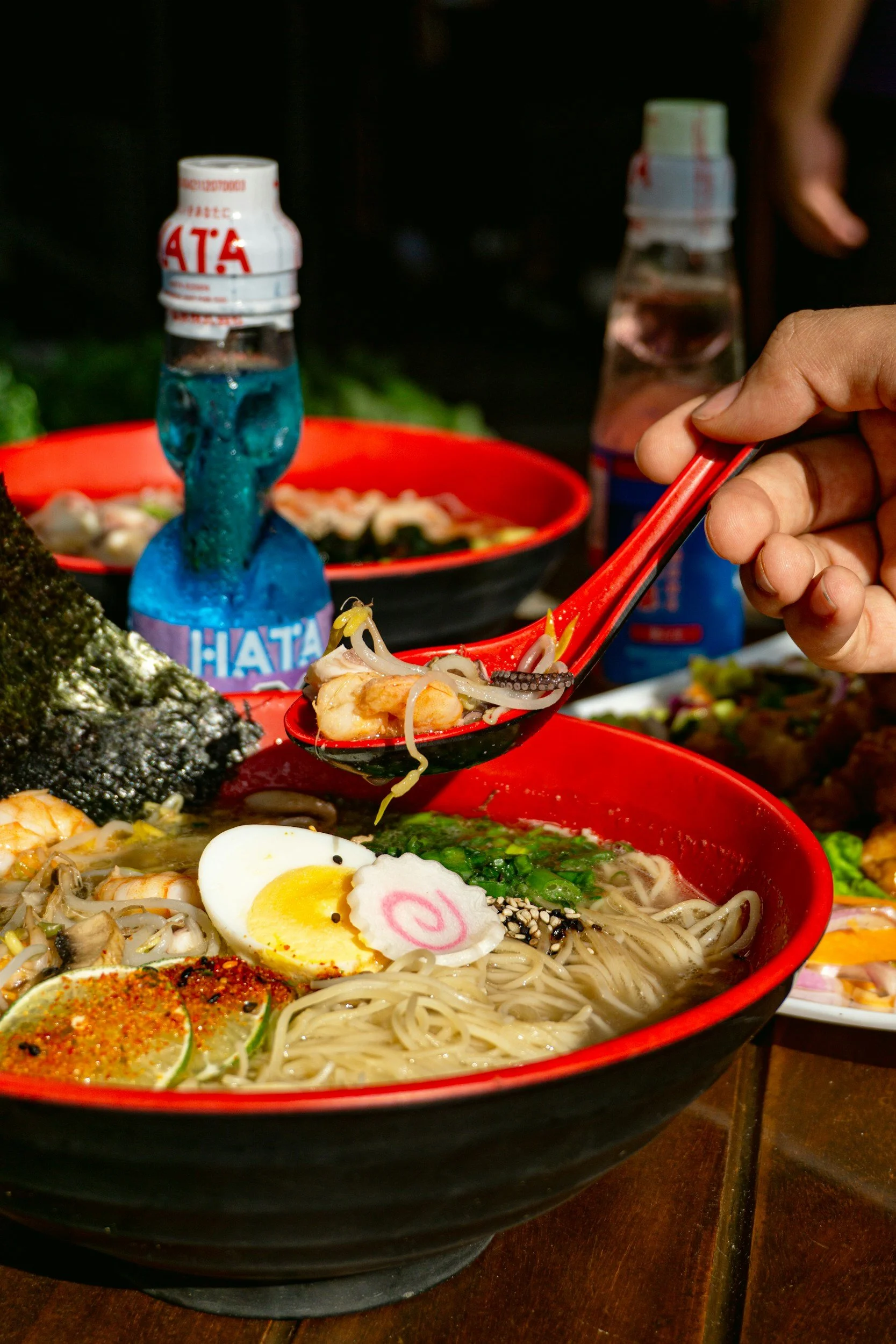 Close-up of a bowl of ramen with sliced boiled egg, narutomaki, green onions, and noodles, with chopsticks holding a piece of food. In the background, there are other bowls of ramen and bottled drinks.