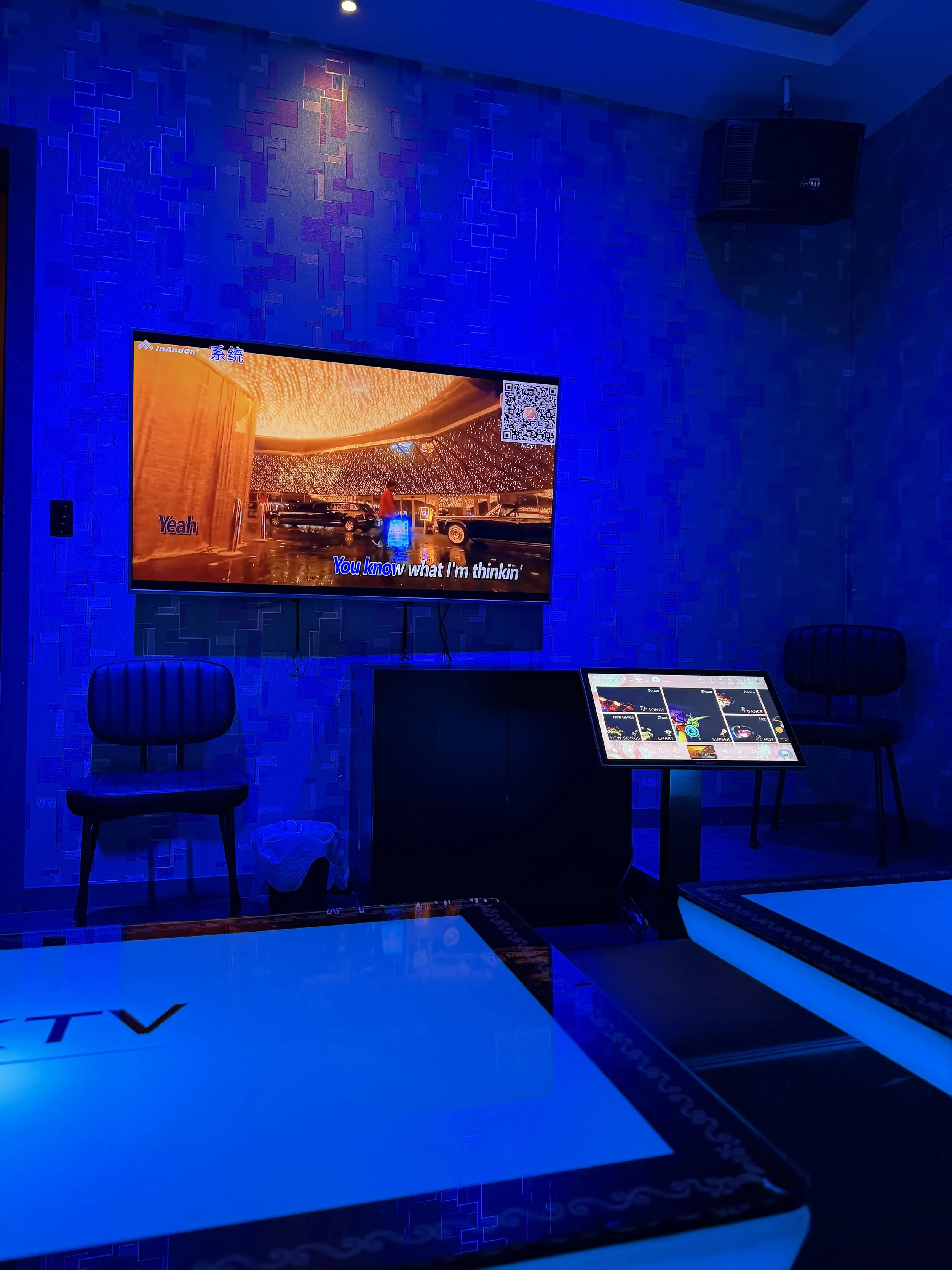 A room with blue lighting, a television screen displaying karaoke lyrics, and a tablet on a stand showing a song selection menu. There are chairs and a table visible, with posters or signs on the table.