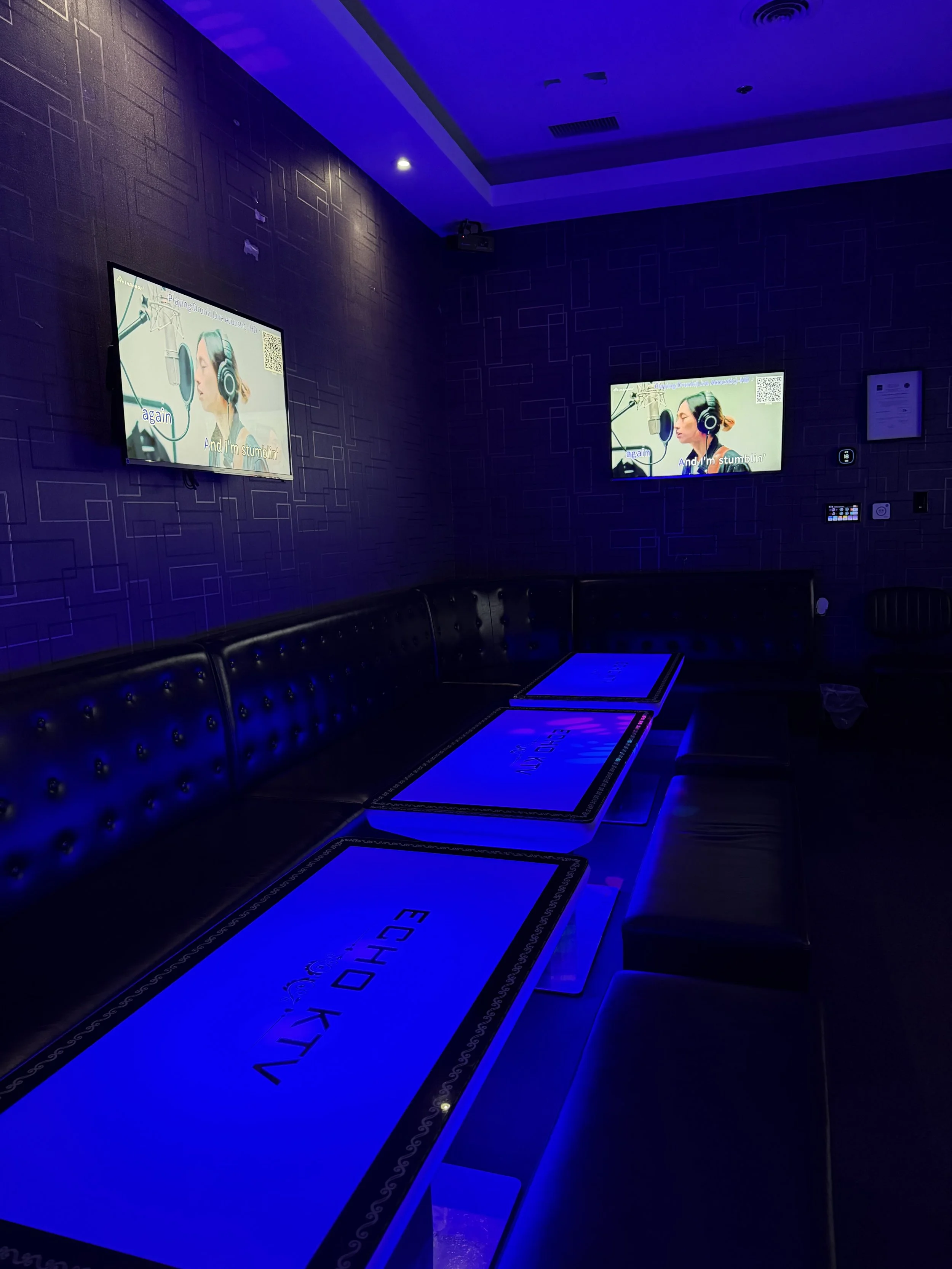 Darkened karaoke room with two large screens displaying lyrics, and a long table with illuminated panels, illuminated by blue lighting.
