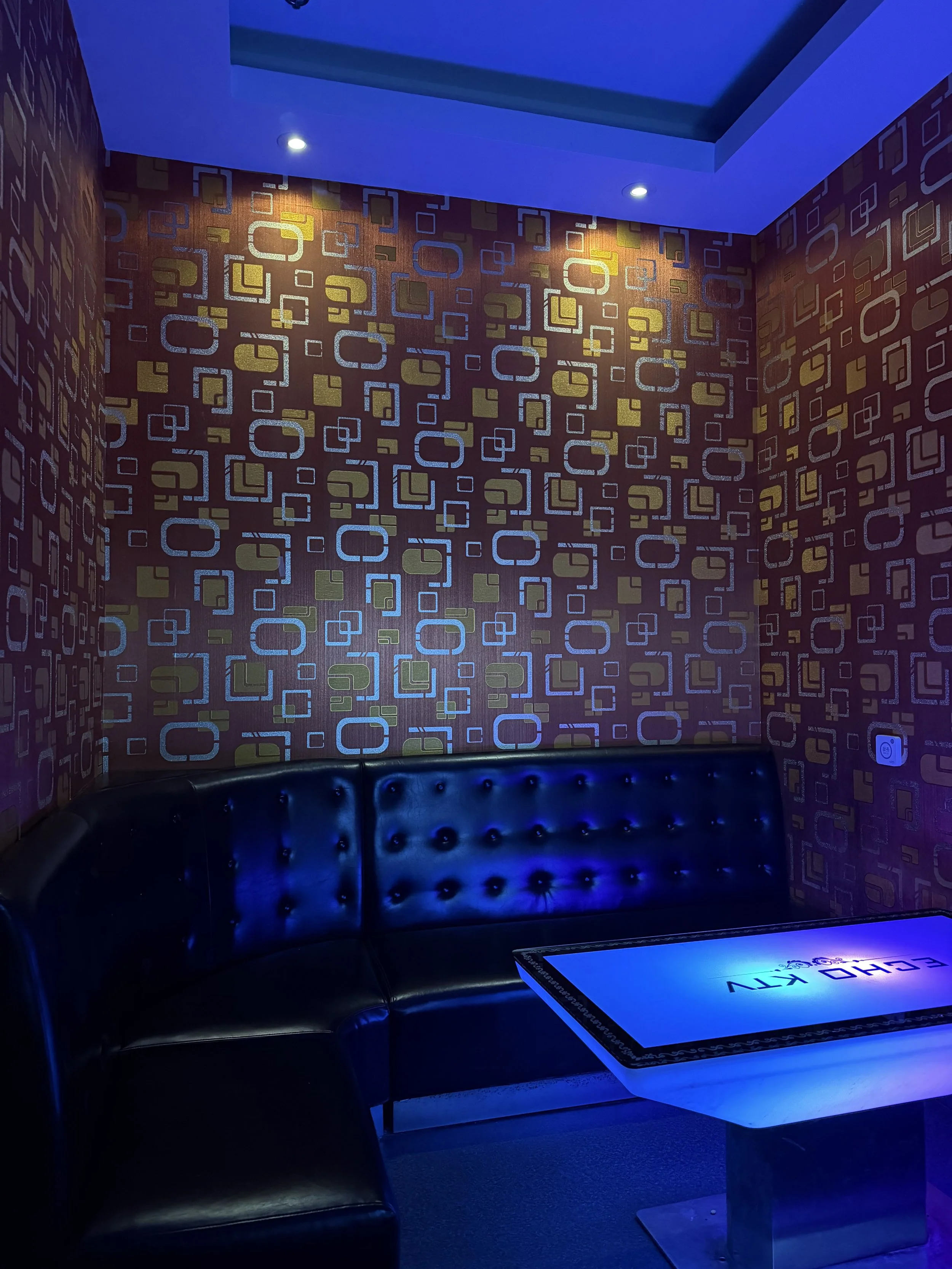 A dimly lit room with a black leather curved sofa, a patterned wallpaper with squares and rectangles, a blue-lit ceiling, and a table with the words "ECHO KEV" illuminated.