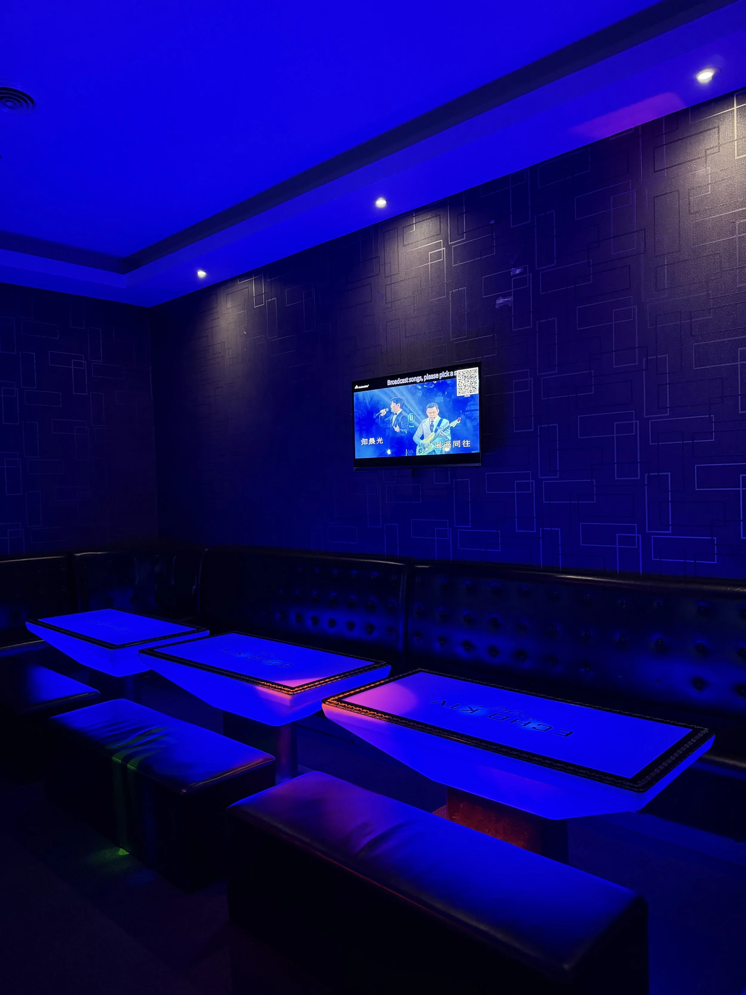 A dimly lit karaoke room with blue and purple lighting, featuring black cushioned seating, white tables, and a wall-mounted television displaying karaoke lyrics and images of two musicians.