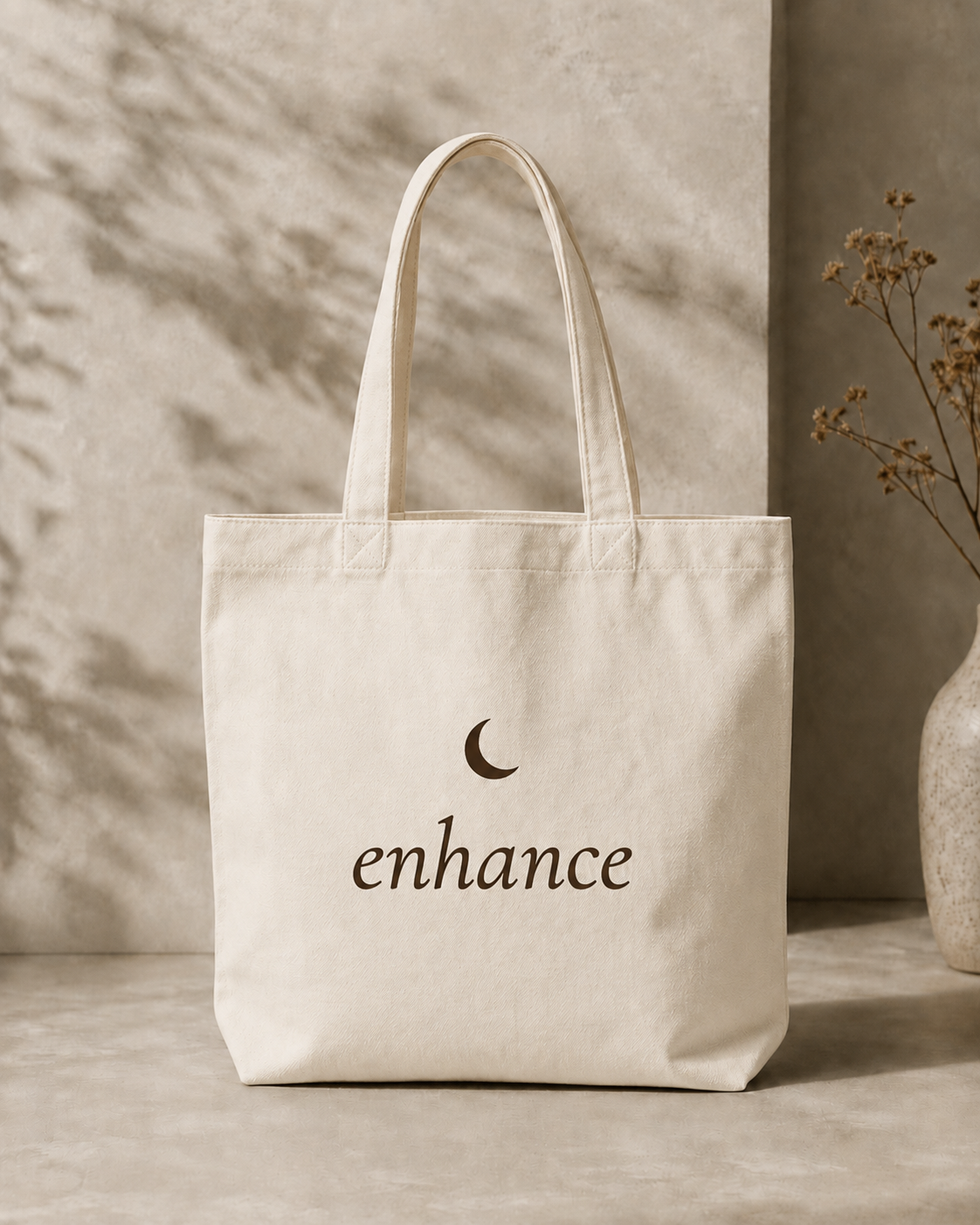 Cream canvas tote bag with the Enhance crescent moon logo, propped beside a vase in soft natural light.