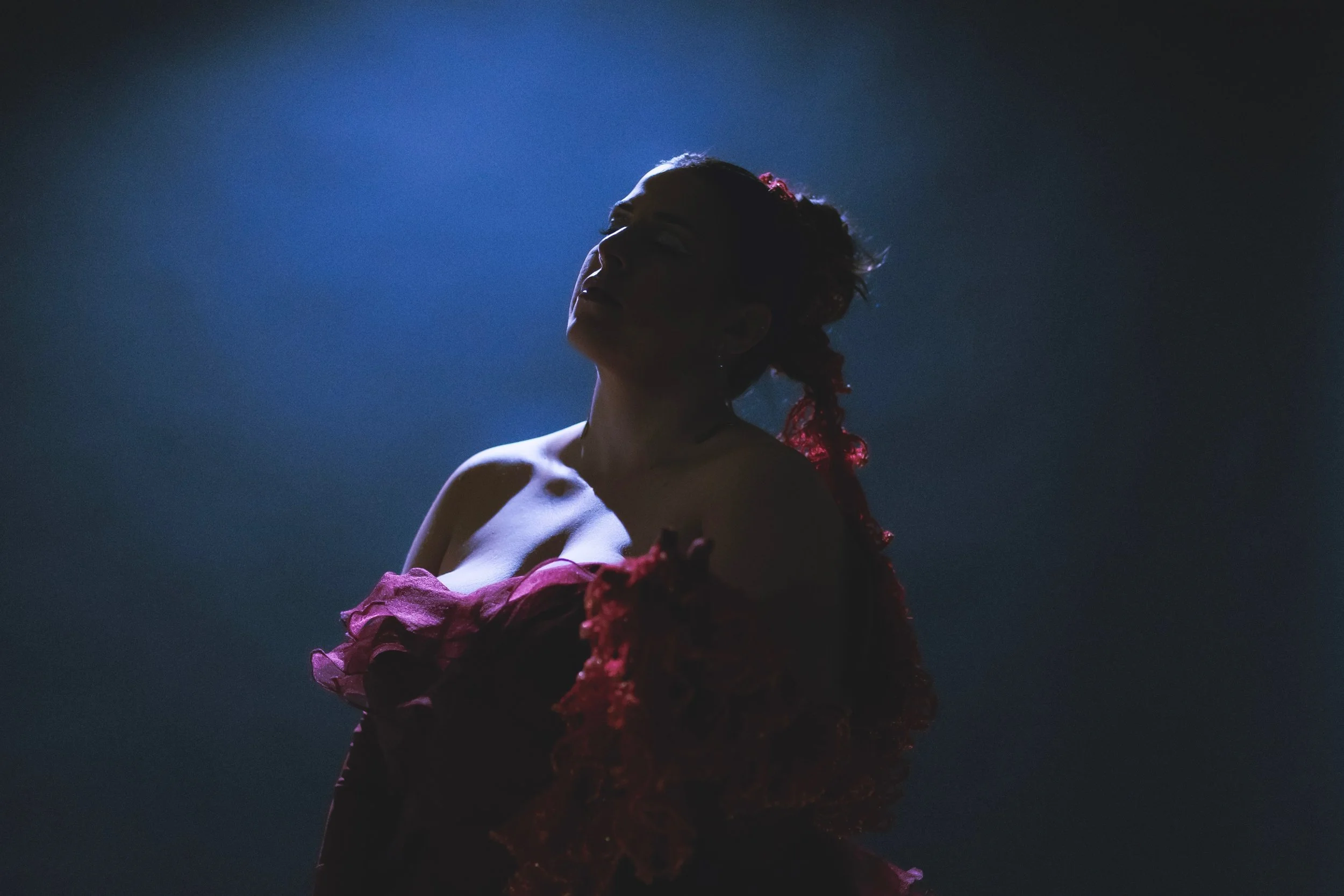 Rnb singer named Taylin Archini with curly hair in profile, wearing a burgundy, ruffled dress, standing in a dark environment illuminated from above with a blue-toned light while she films her music video.
