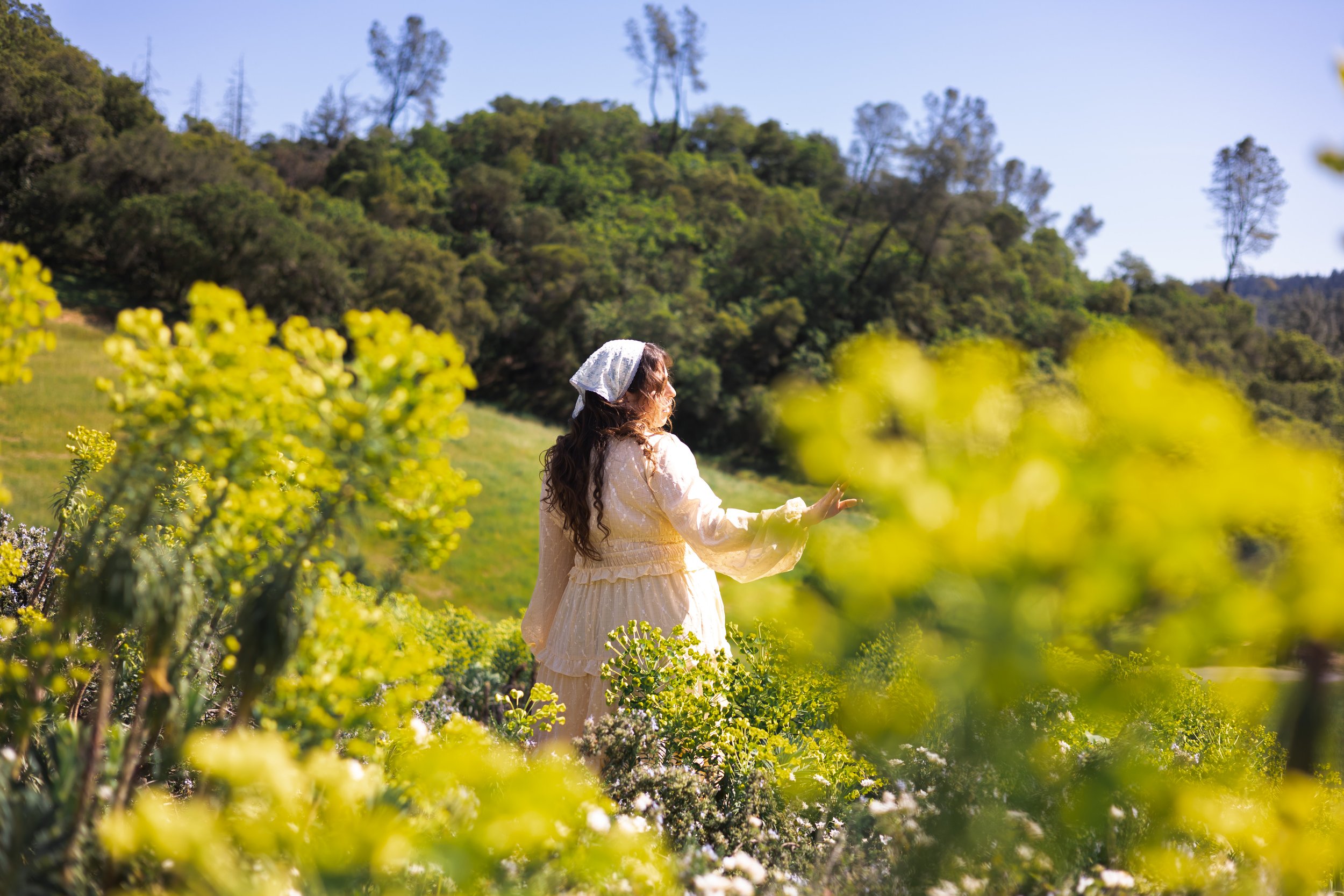 Rnb singer named Taylin Archini in a cream-colored dress and white headscarf standing in a lush green field with yellow flowers, surrounded by trees and hillside under a clear blue sky for her music video to the song Leave Me Alone.