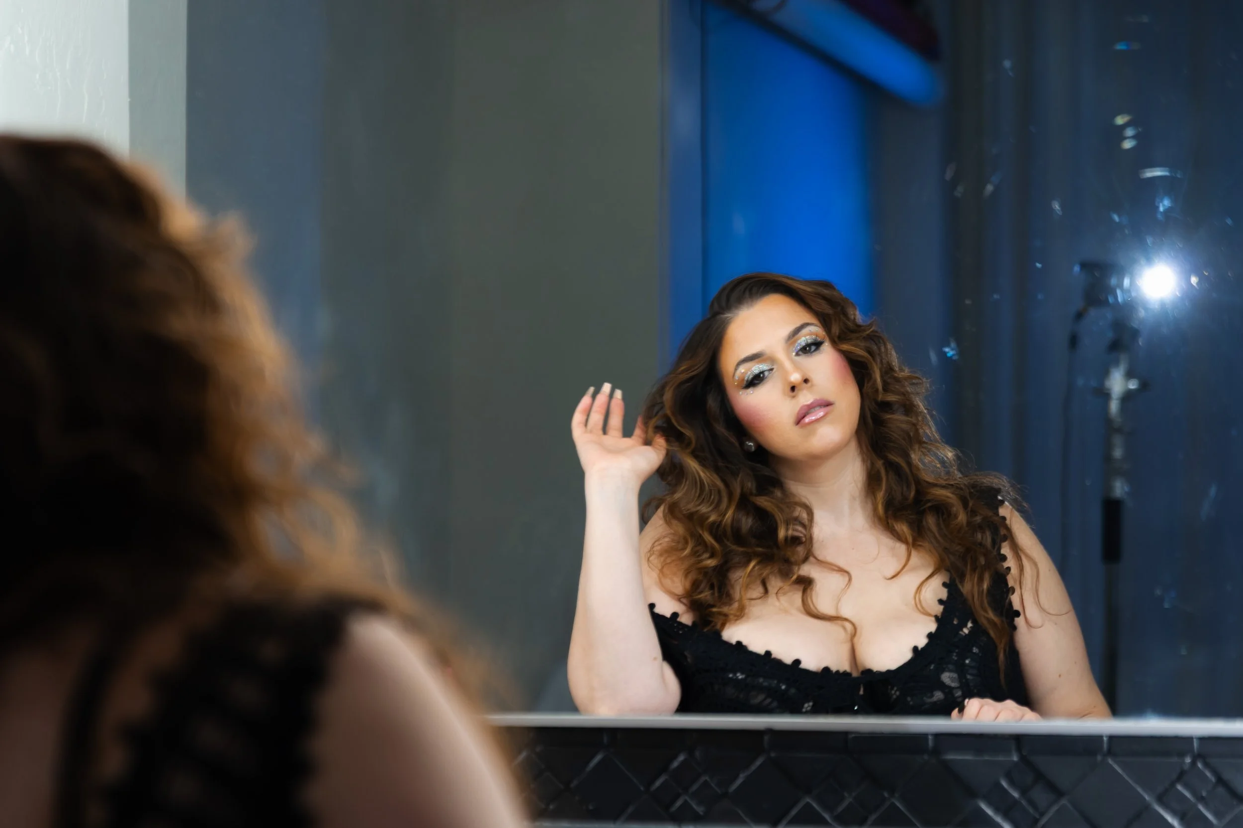 Rnb singer named Taylin Archini with long curly brown hair and glamorous makeup, including sparkly eye makeup, wearing a black lace dress, looking at herself in a mirror as she adjusts her hair during a photoshoot for her EP cover.