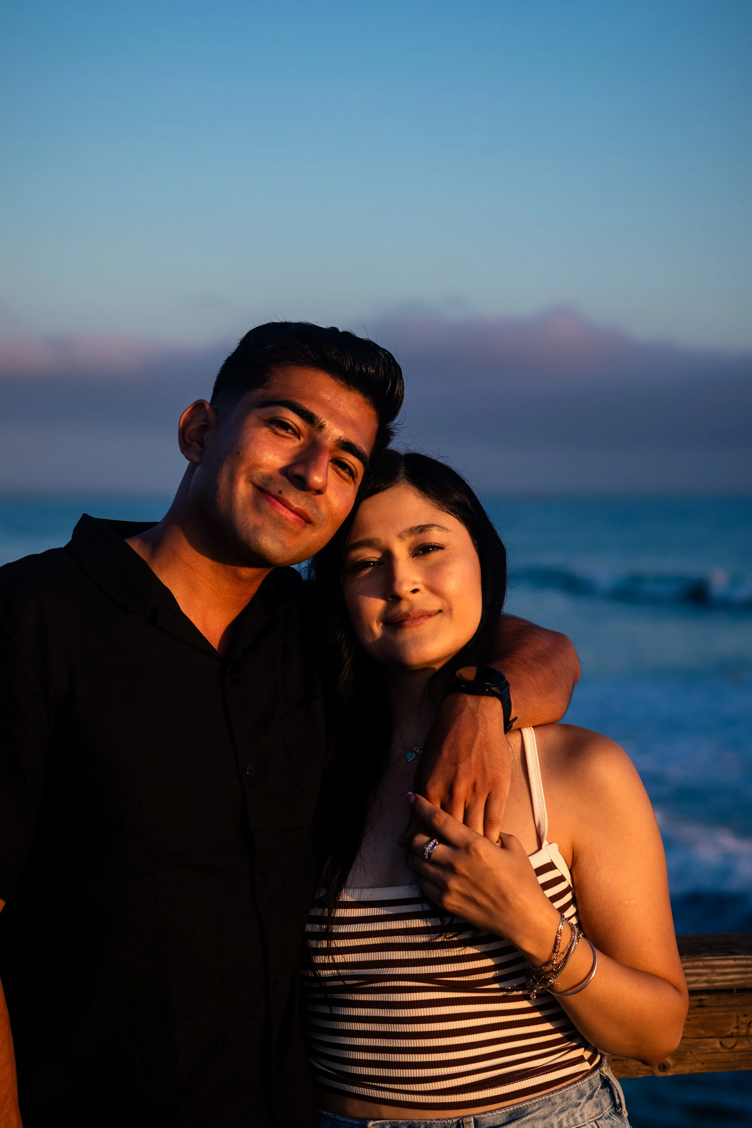 A young couple smiling and embracing each other on a beach at sunset, with the ocean and sky in the background.