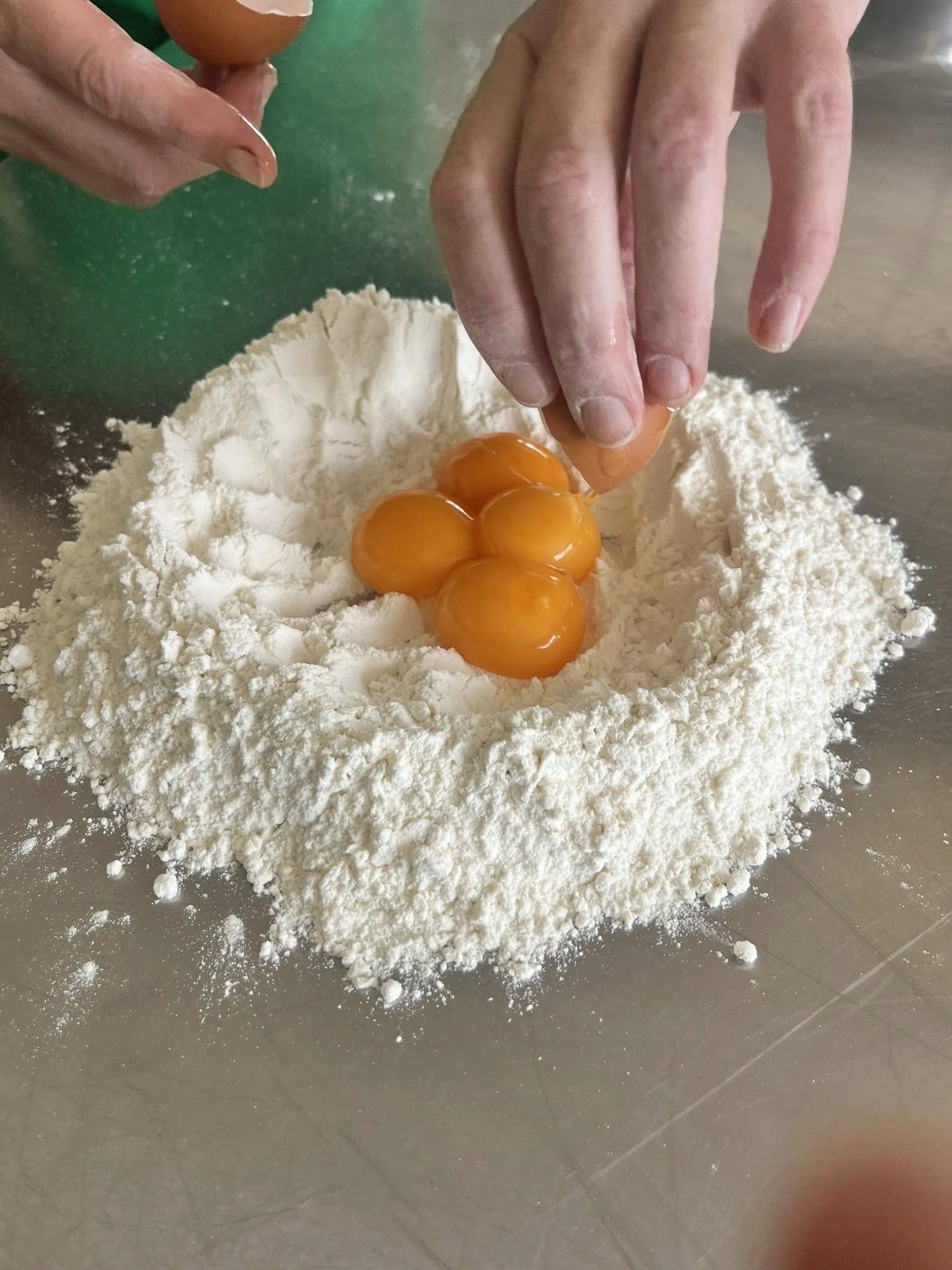 A person cracking eggs into a pile of flour to make fresh pasta on a stainless steel surface.