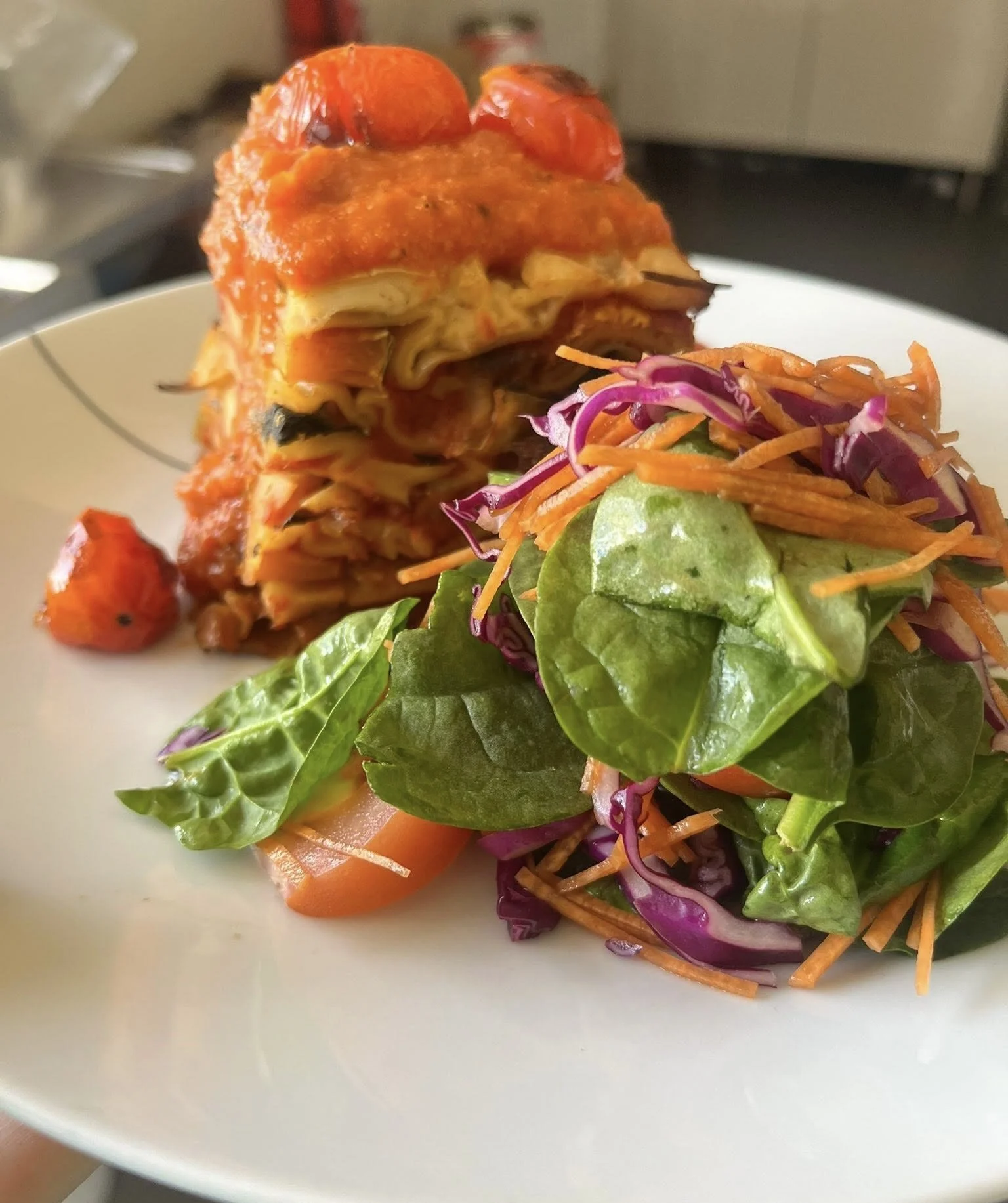 A plate with a portion of lasagna topped with tomato chunks and a side of mixed greens salad with shredded carrots and purple cabbage.