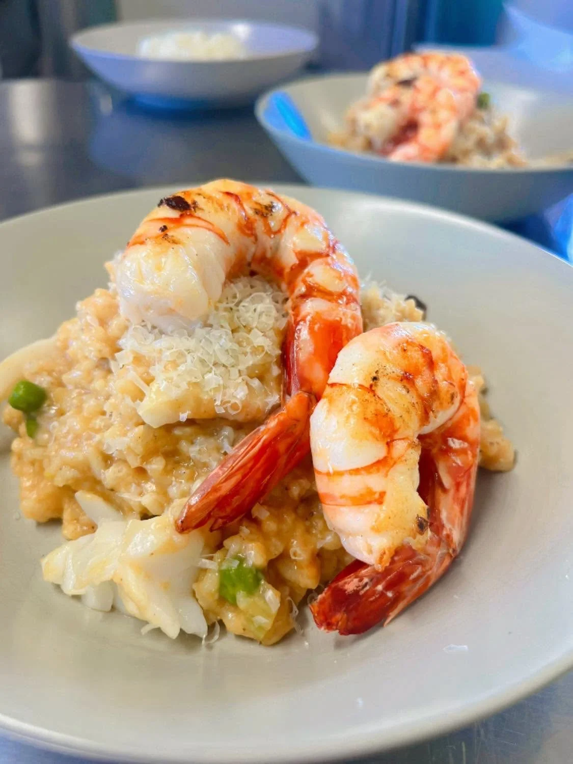Shrimp and rice dish garnished with grated cheese on a white plate. In the background, two more plates of the same dish and a bowl of white sauce or cheese.