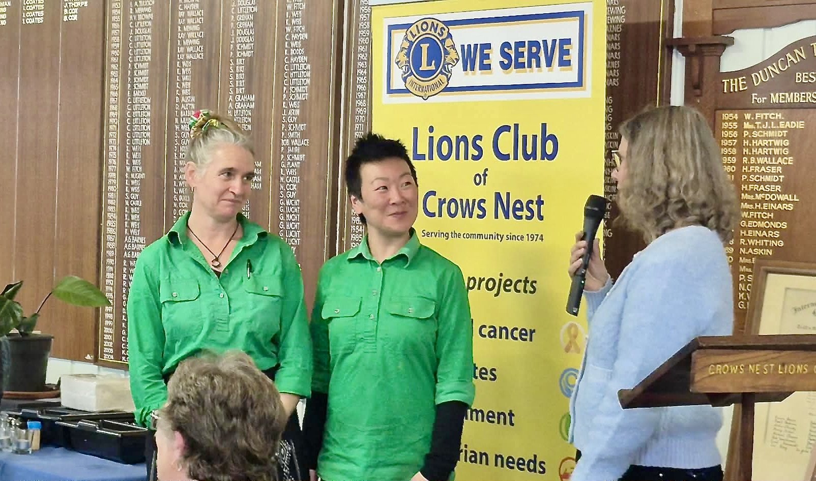 Vanessa and Kat being congratulated by a Lions Club commitee member with a yellow Lions Club banner in the background.