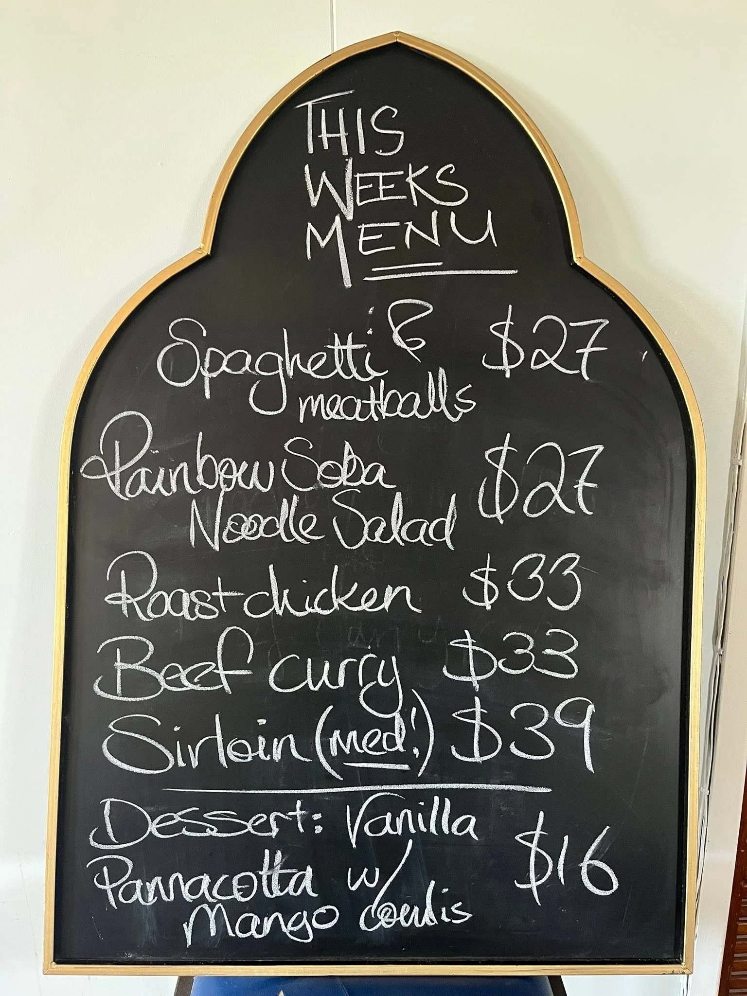 A black chalkboard menu with a golden border listing weekly menu items and prices, written in white chalk.