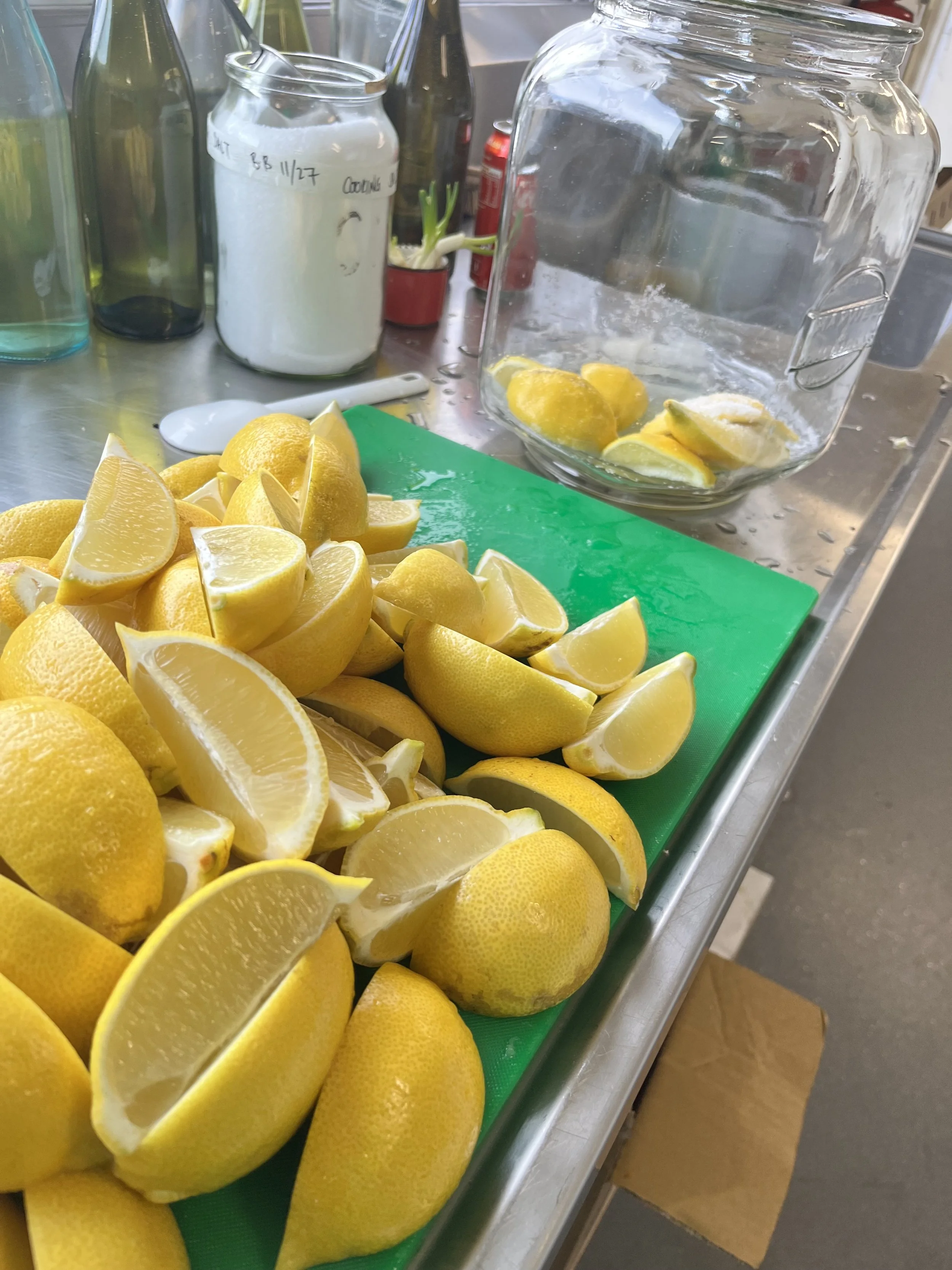 A large pile of freshly cut lemons on a green cutting board on a metal countertop, with a glass jar filled with lemon slices and ice, and various bottles and containers in the background.