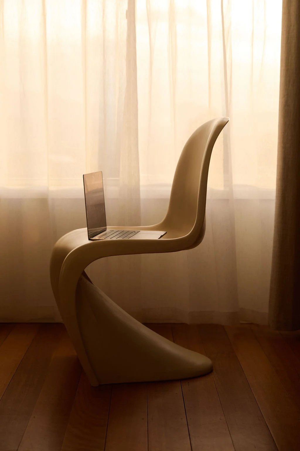 A sleek, curved cream-coloured Panton chair sits in profile with an open laptop balanced on its seat, set against sheer curtains lit by soft natural light. The chair's sculptural S-shaped form casts a shadow across the timber floorboards.
