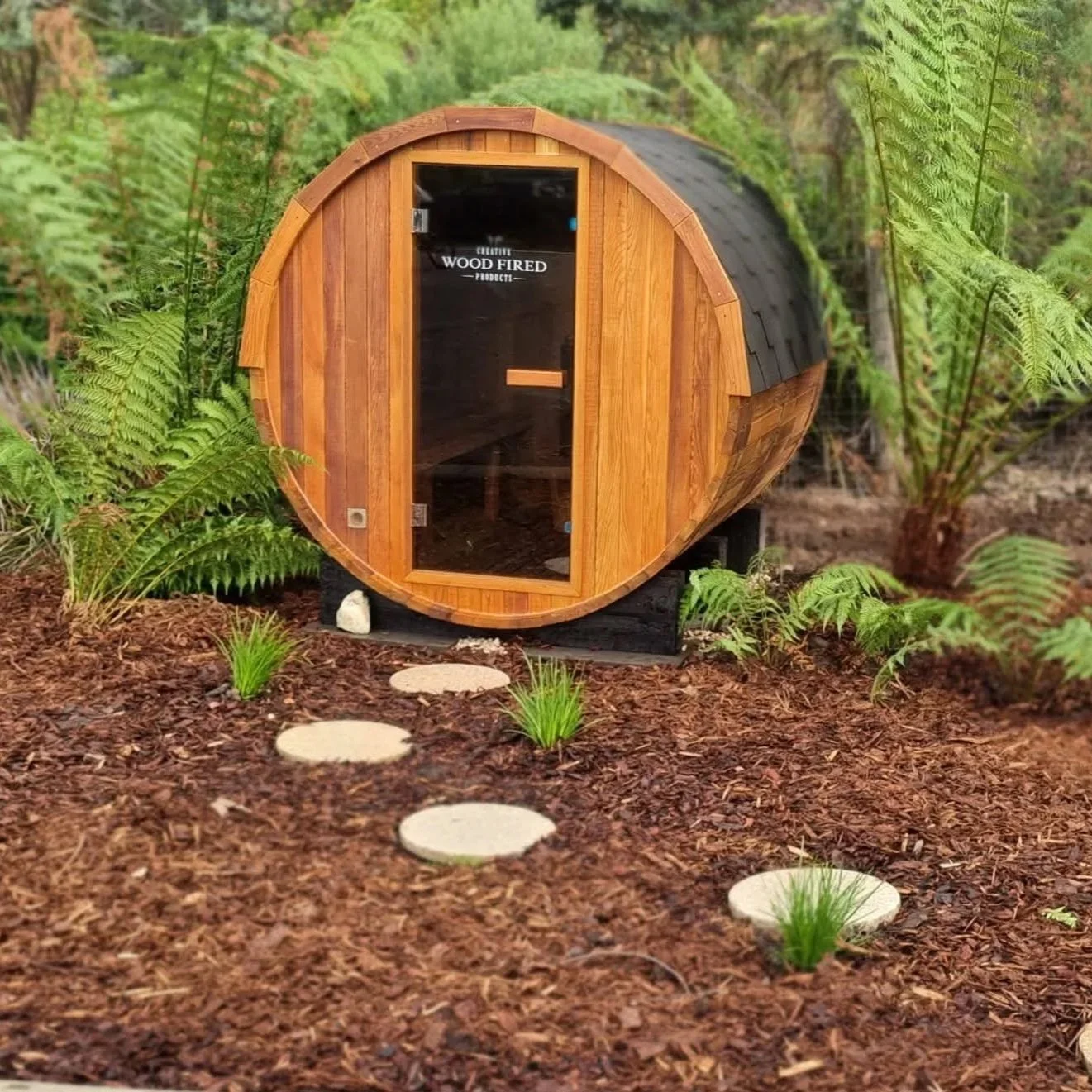 A small wooden barrel sauna with a glass door, set in a garden surrounded by ferns and small plants, with a stepping stone pathway leading to it.