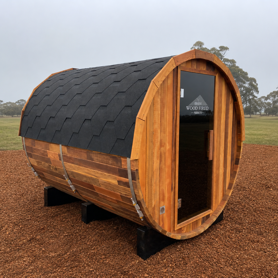 A small wooden barrel-shaped sauna with a glass door, black shingle roof, and wooden siding, placed on a gravel surface in an open field with trees in the background.