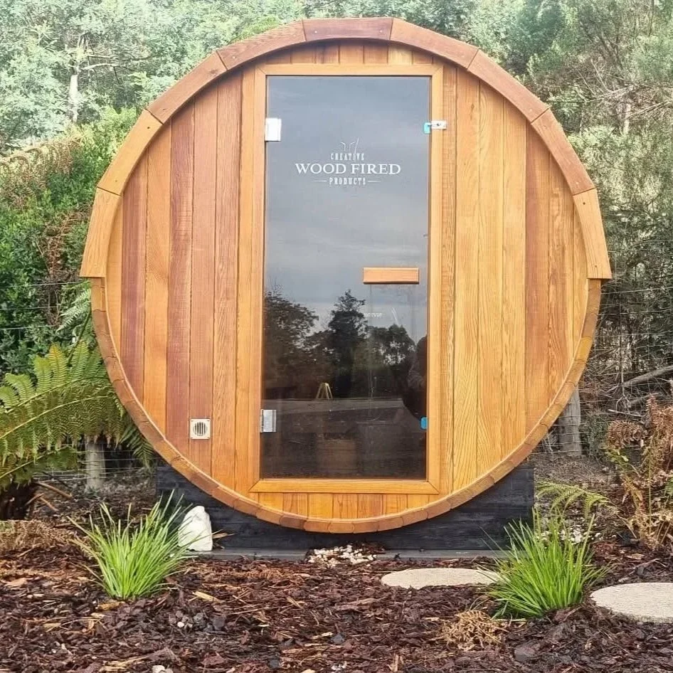 Wood-fired sauna with a round shape and a glass door, surrounded by greenery and plants.