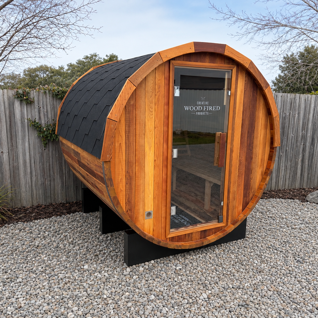 Small, barrel-shaped wooden sauna with a glass door and dark shingle roof, situated outdoors on a gravel surface with a wooden fence and trees in the background.