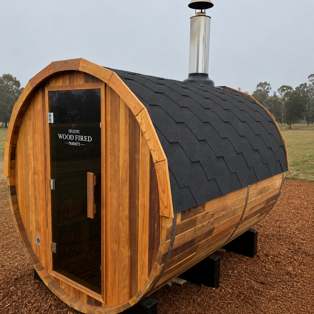 A wooden outdoor sauna with a curved shape, black shingle roof, a metal chimney, and a glass door with the words "Creative Wood Fired Products" on it, situated on a grassy field.