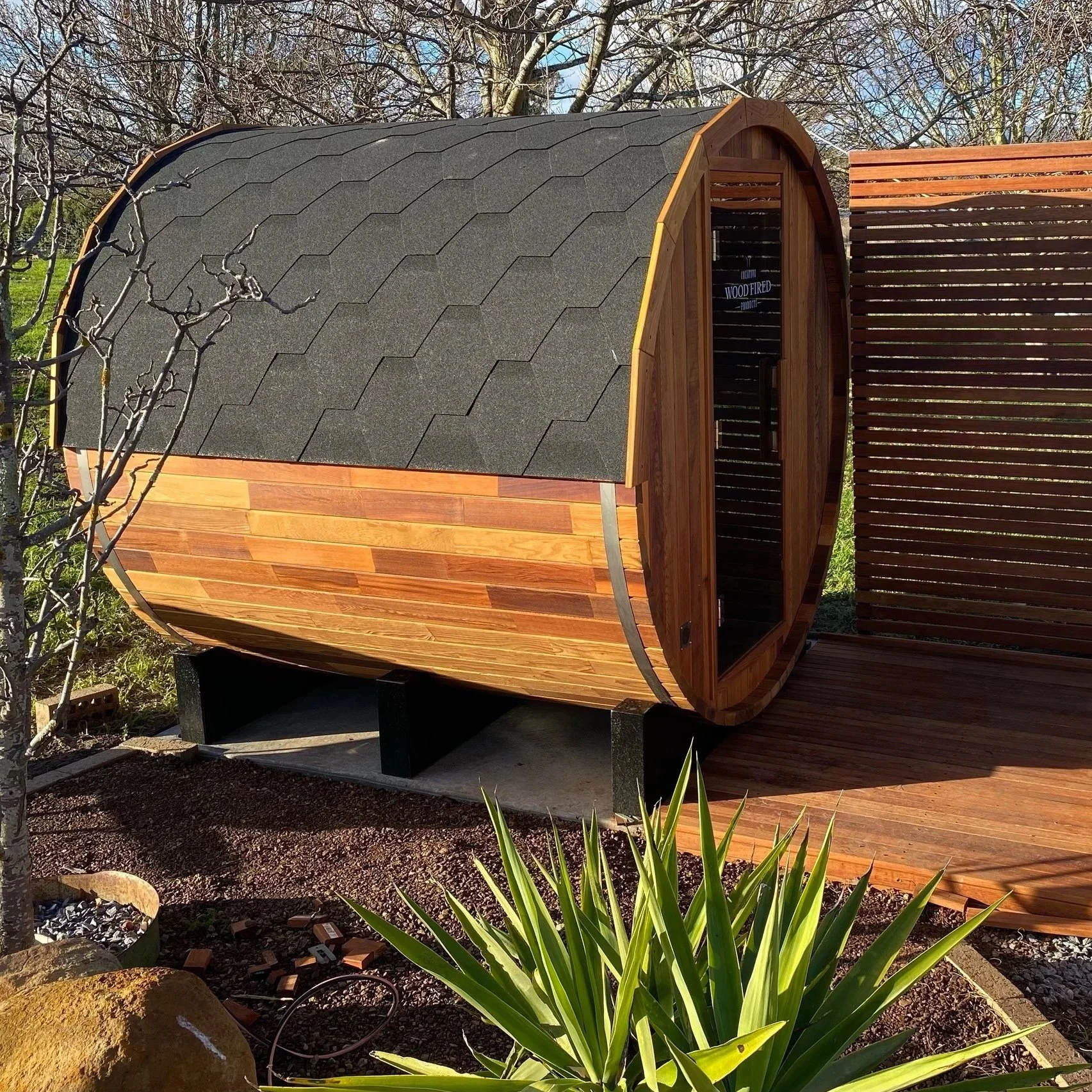 A small, barrel-shaped wooden sauna with a black shingled roof and a small door, set outdoors next to a wooden deck and surrounded by plants and gravel, with a background of leafless trees.