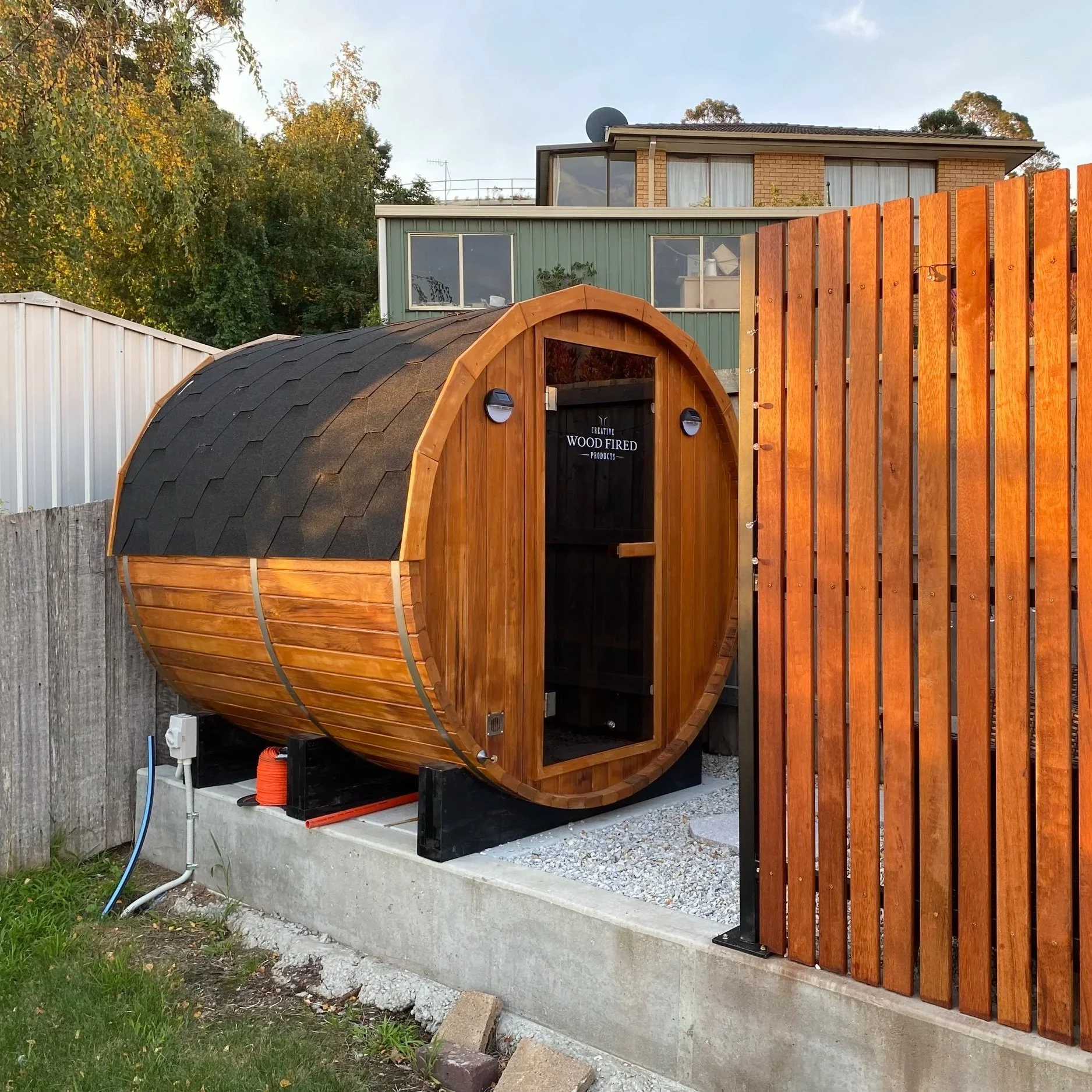 A small wood-fired oven with a curved, shingle roof and a black door, situated on a concrete foundation with a wooden fence on the right side and a residential house with large windows in the background.