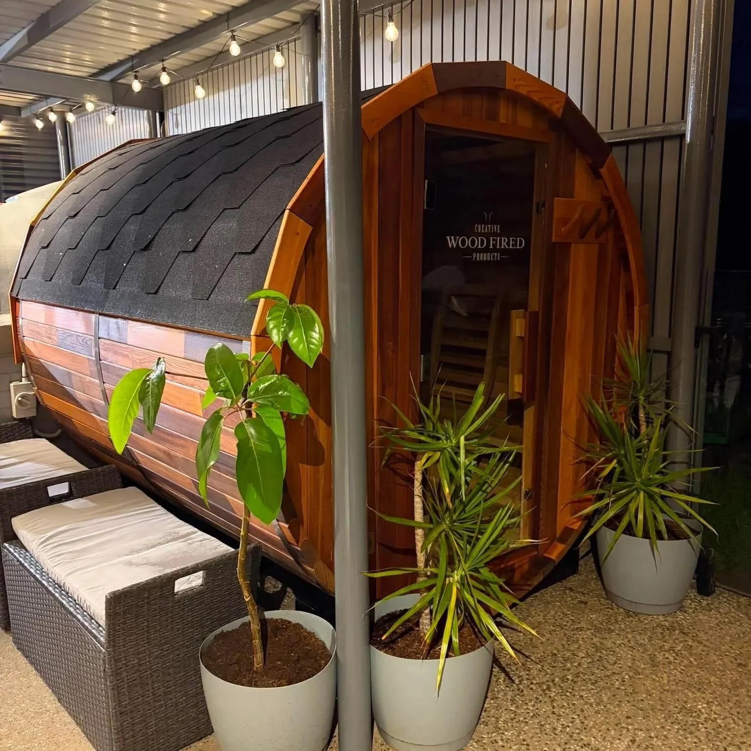 A small wooden sauna surrounded by green potted plants, with a bench outside and string lights hanging overhead.