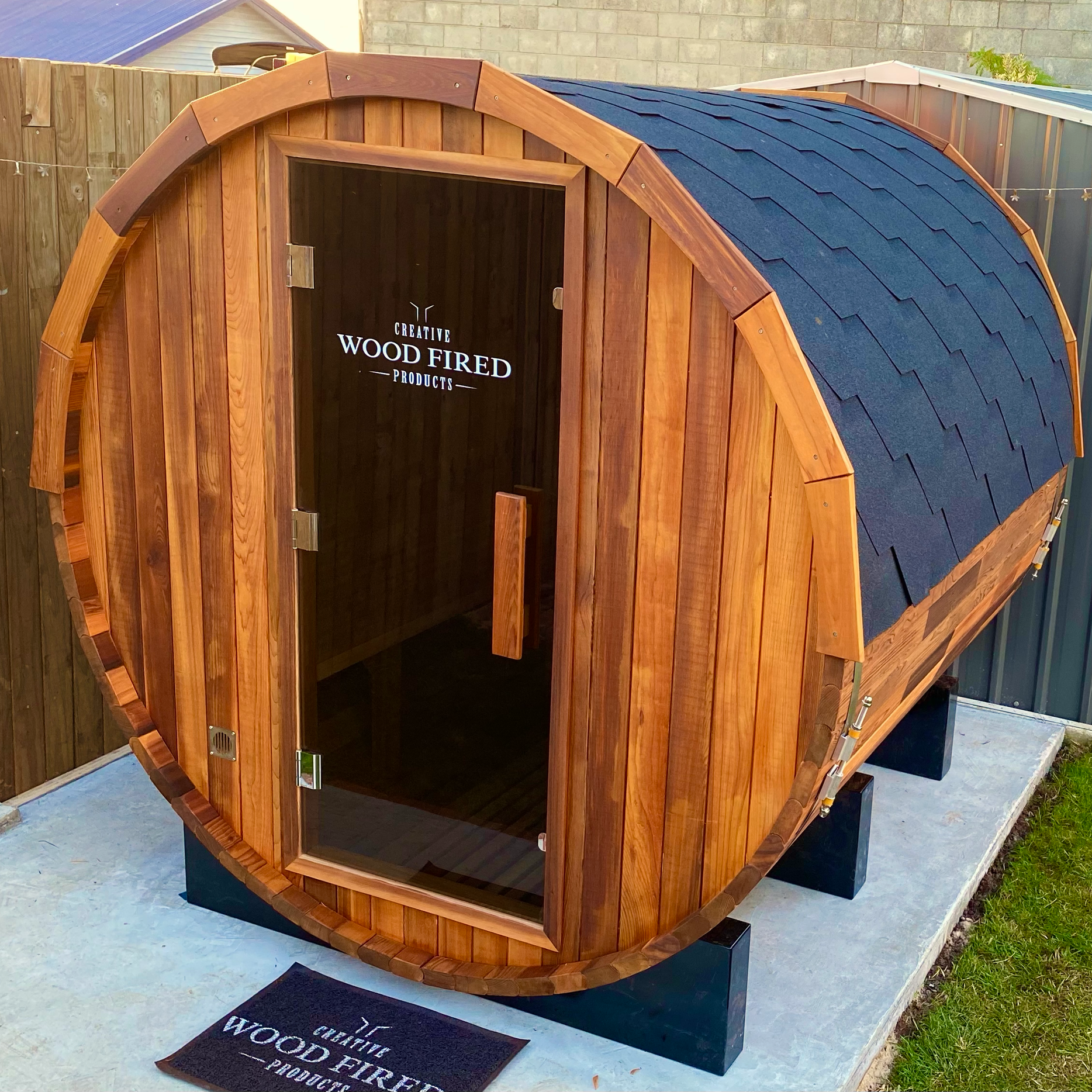 A wooden outdoor sauna with a rounded roof covered in blue shingles, situated on a concrete patio with a black mat in front that reads "Wood Fired". The sauna has a glass door and an interior with wooden paneling. A small black vent is visible at the