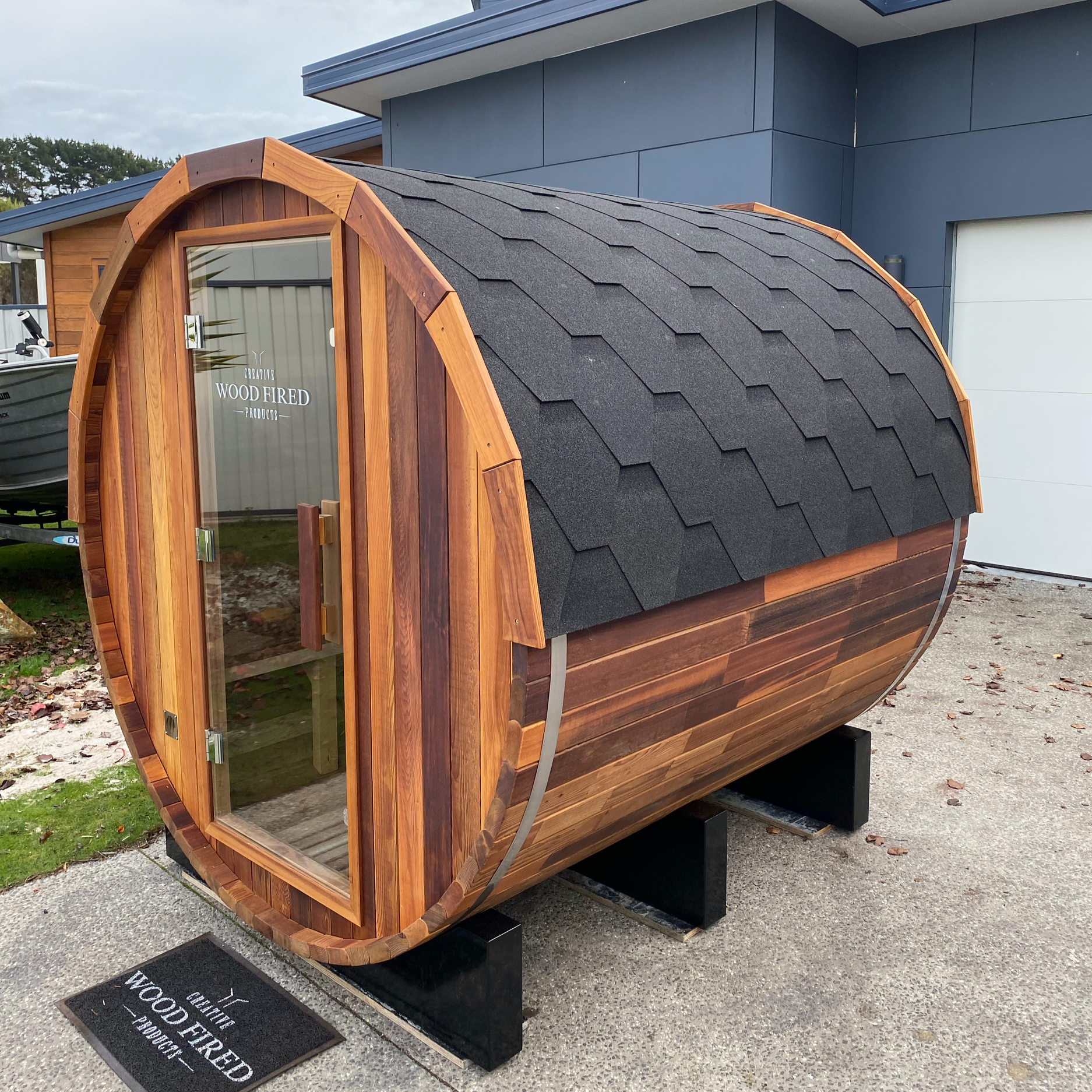 A small wooden structure with a rounded roof covered in black shingles, glass door, and a sign that reads "Creative Wood Fired Products." It is situated outdoors on a paved surface, with a modern house and a boat in the background.