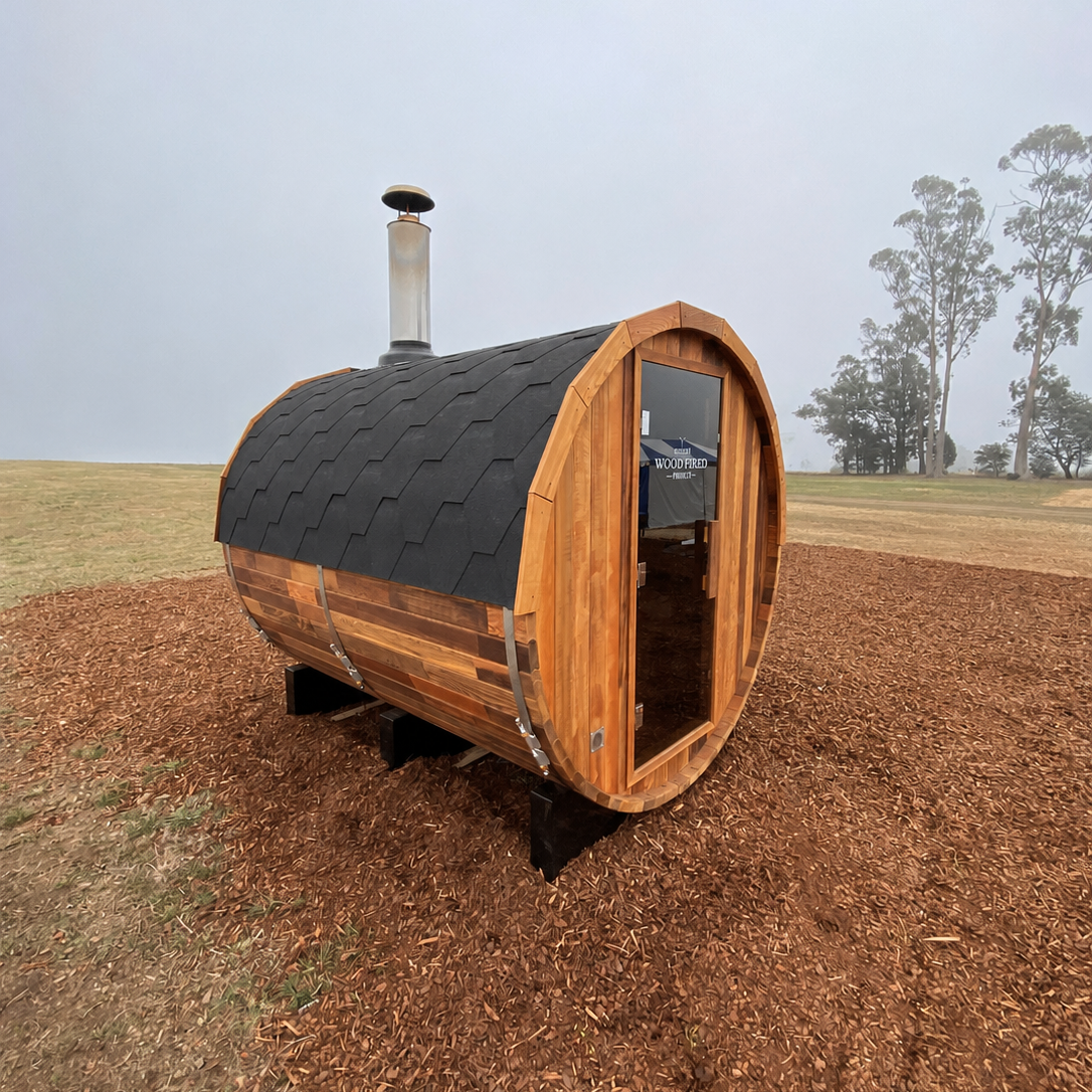 Small wooden barrel-shaped outdoor oven with black shingle roof and a chimney, situated on wood blocks amid a grassy field with trees in the background.