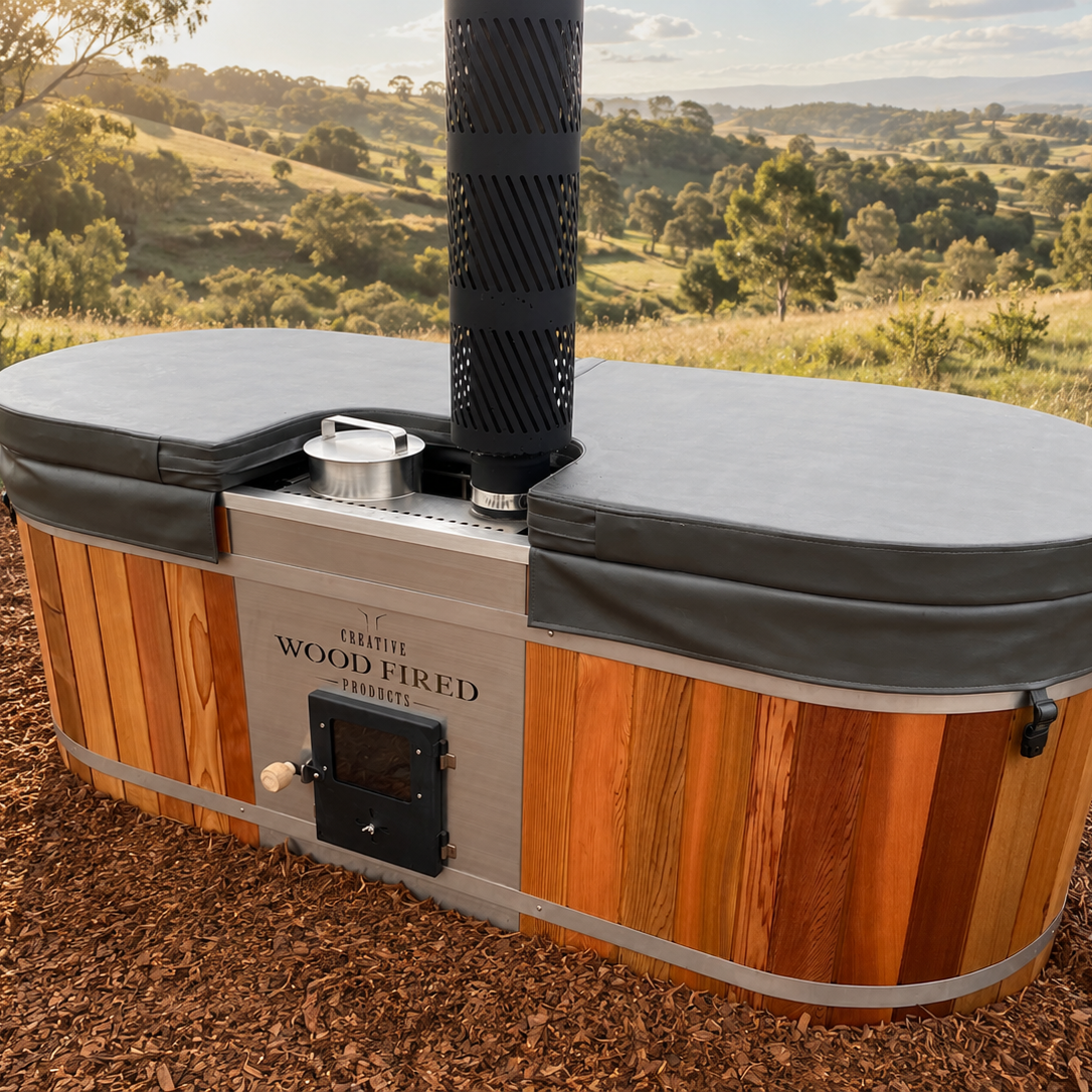 A wooden hot tub with a black stove pipe chimney, set outdoors on dirt ground with a scenic landscape of rolling hills and trees in the background.