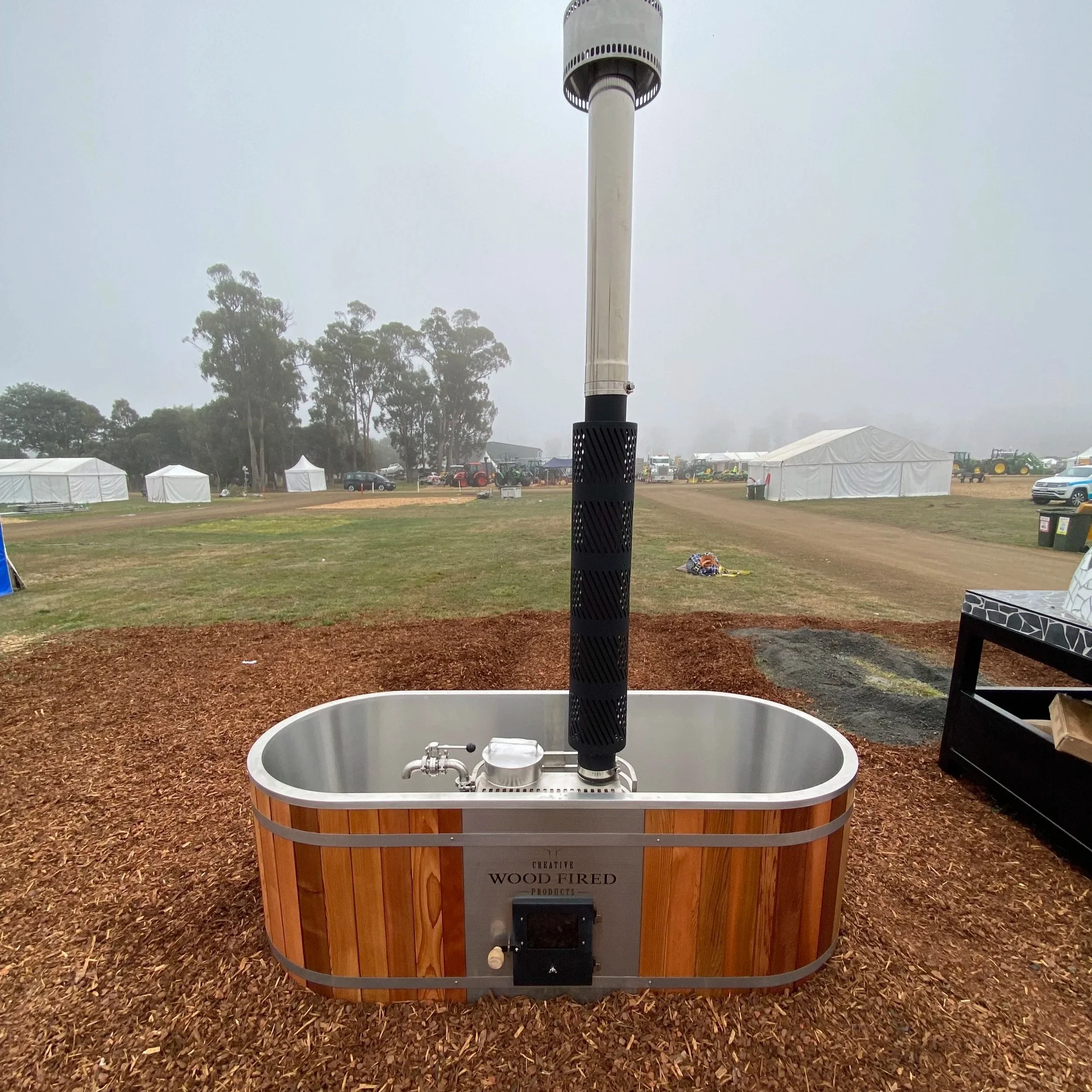 A wooden outdoor fire pit with a metal chimney pipe on top, labeled "Creative Wood Fired Products" in front of a foggy outdoor setting with tents, trees, and tractor equipment in the background.