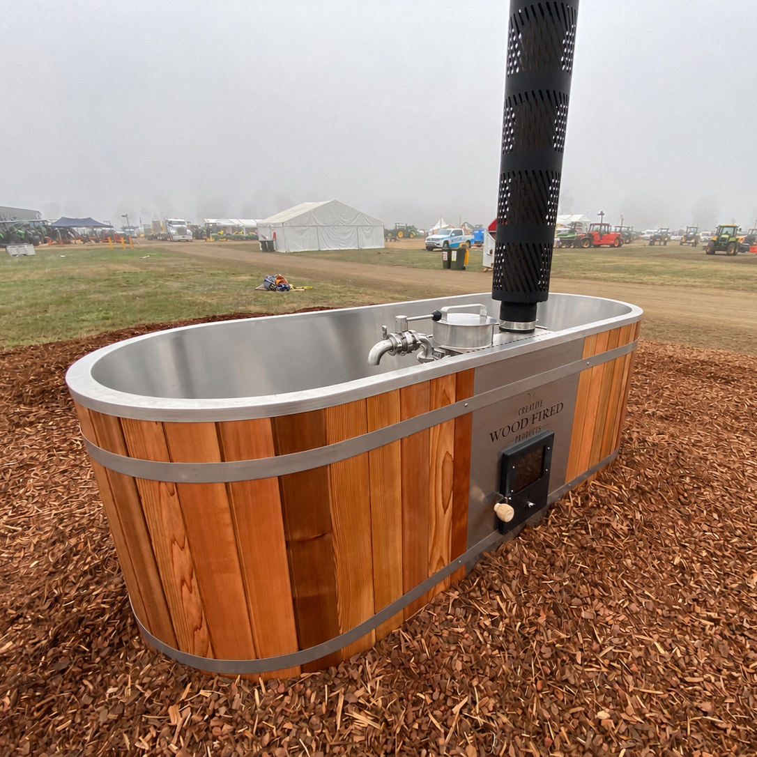 A metallic, wood-finished outdoor bathtub with a black chimney and plumbing fixtures, situated on wood chips in a foggy outdoor event space with tents and vehicles in the background.