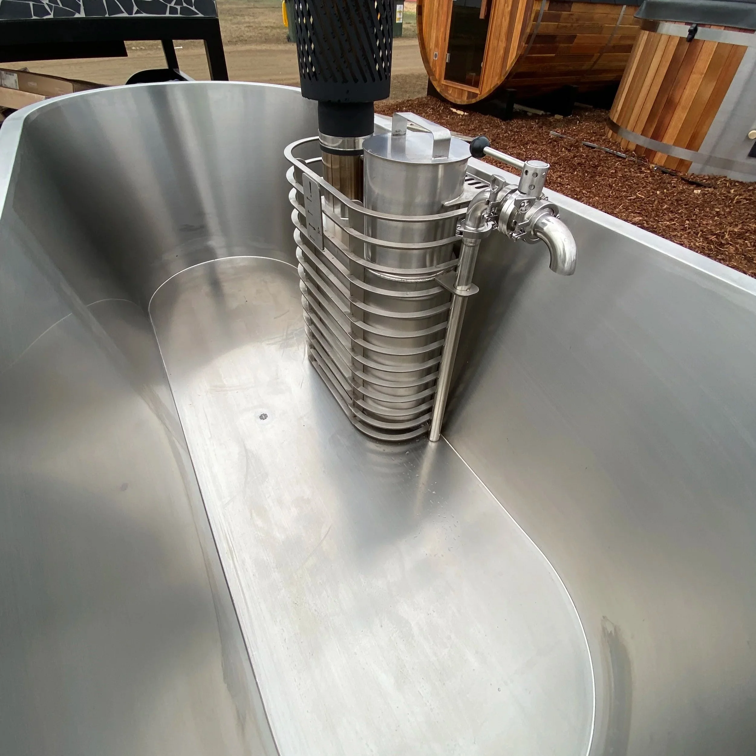 A stainless steel insert for wood fired hot tub, outdoors on a wooden platform with wood chips, barrels, and outdoor structures in the background.
