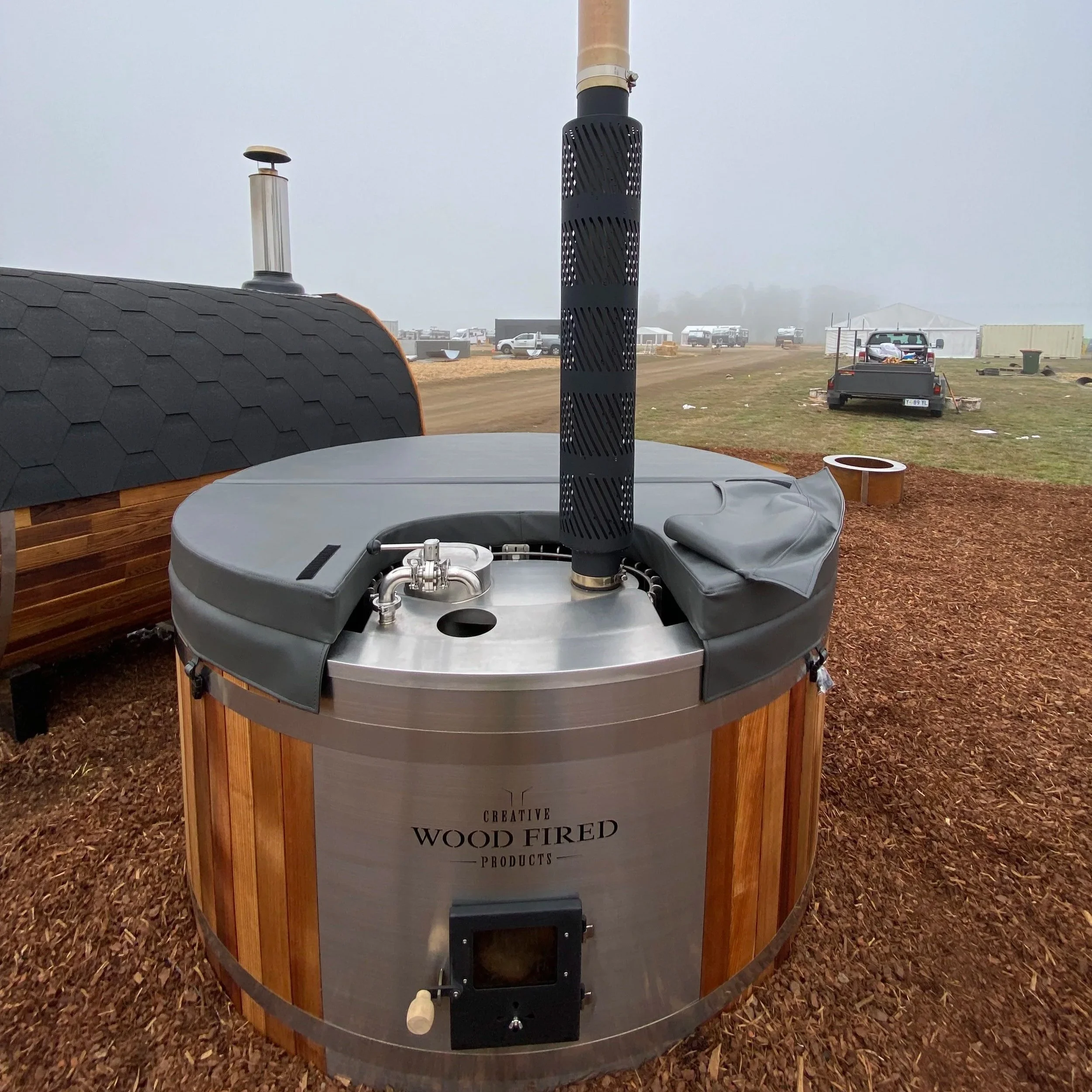 A wood-fired pizza oven with a black chimney and a round aluminum body, set outdoors on a bed of mulch, with a sign that reads 'Creative Wood Fired Products'.
