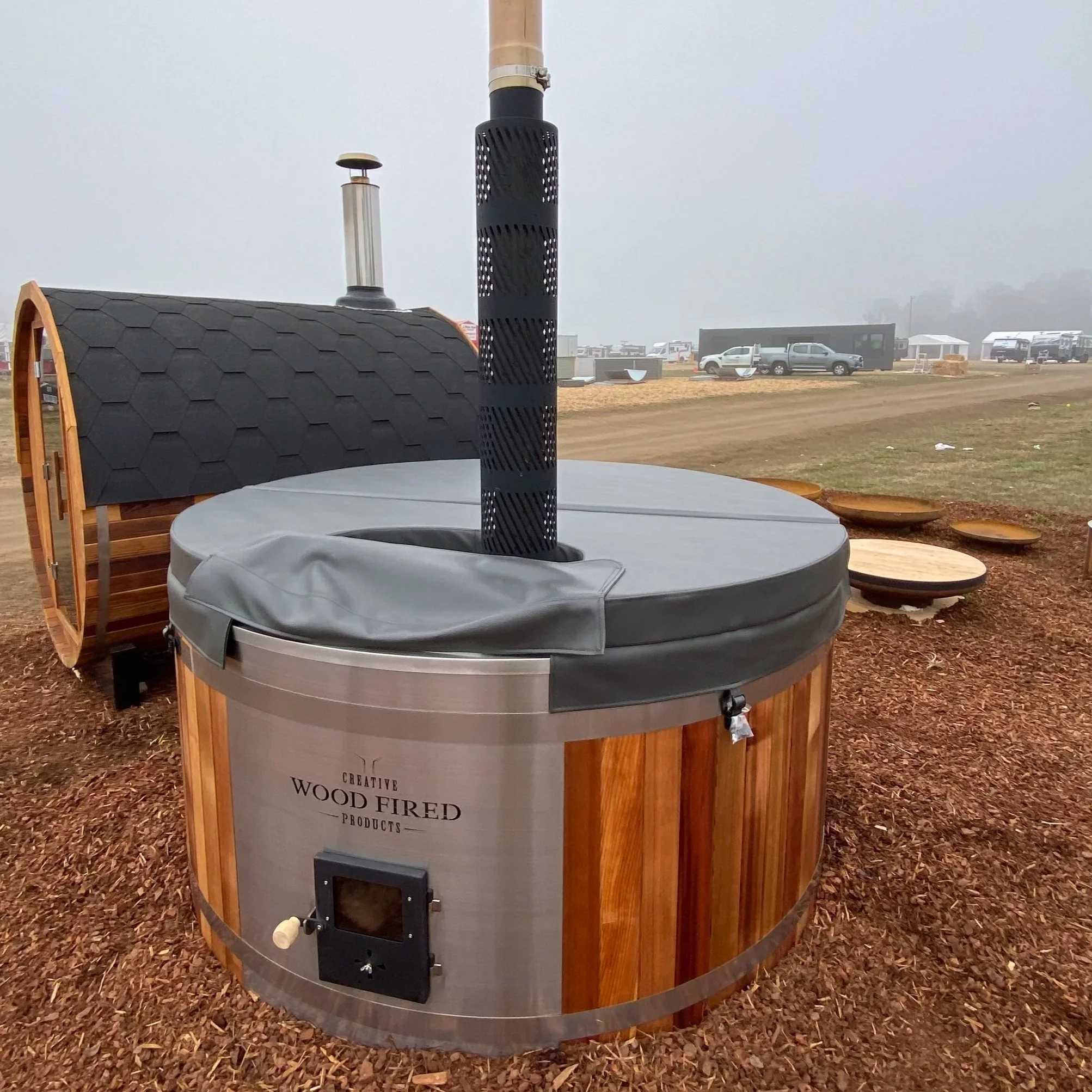 A large wood-fired oven with a rounded metal body and wooden panels, situated outdoors on a ground covered with wood chips. In the background, there are parked vehicles, a black building, and foggy weather, suggesting a rural or festival setting.
