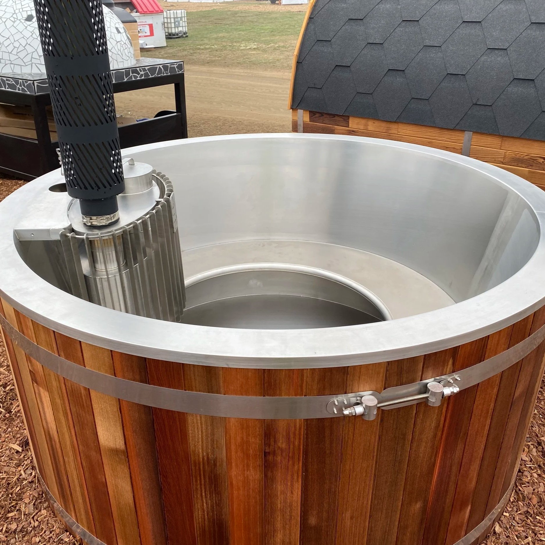 A large outdoor hot tub with a wood exterior, metal trim, and an inset seating area. Part of a black chimney or stovepipe is visible on the left, with a black metal grill at the top. 