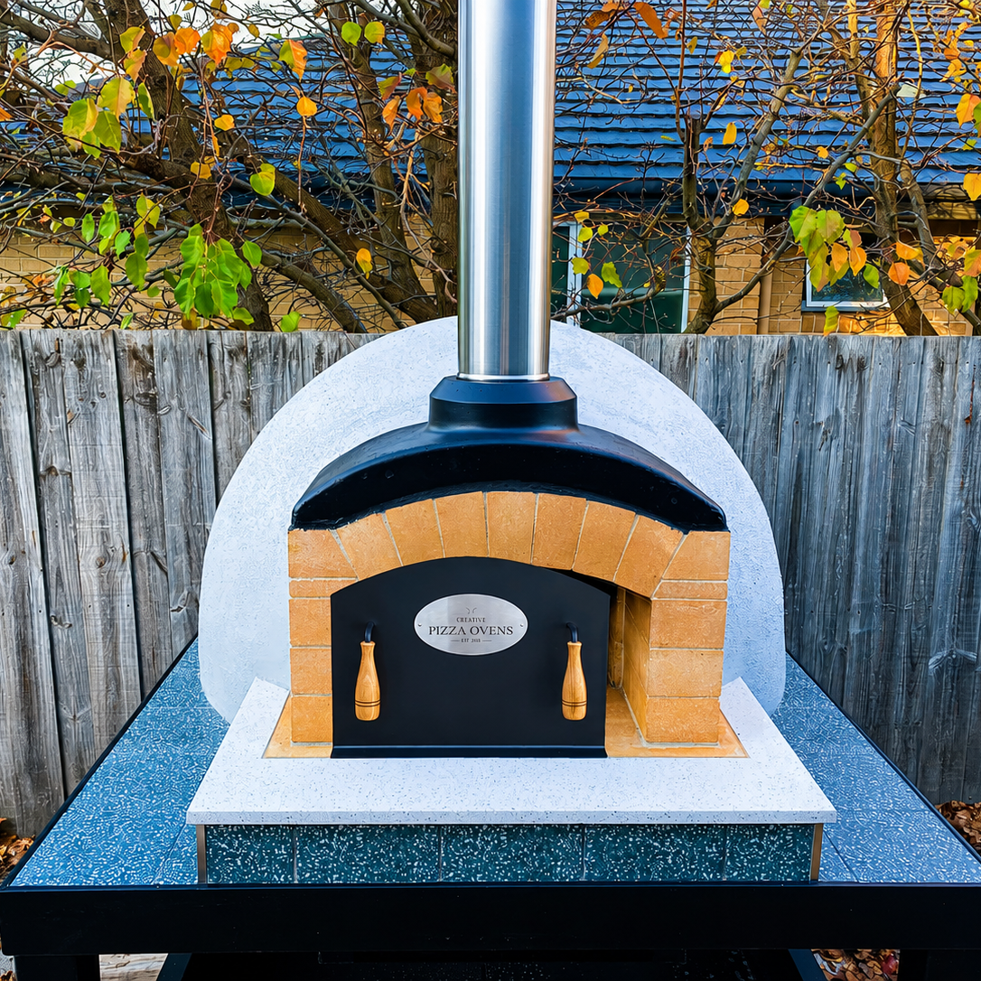 A brick pizza oven with a metal chimney, mounted on a speckled countertop, with a sign that says 'Creative Pizza Ovens'.