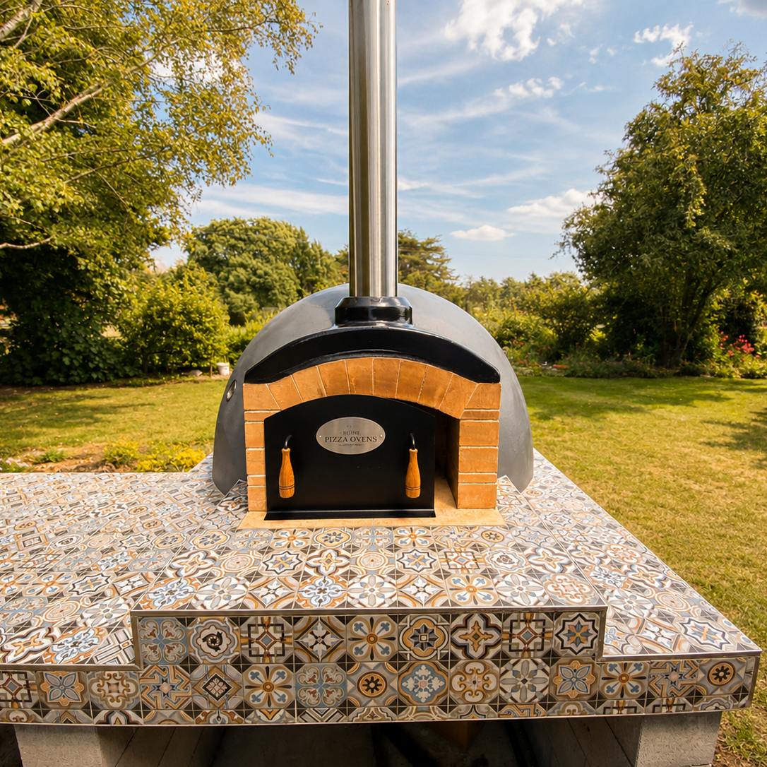 An outdoor pizza oven with a colorful tile countertop on a table, set in a garden with green trees and grass, under a blue sky with some clouds.