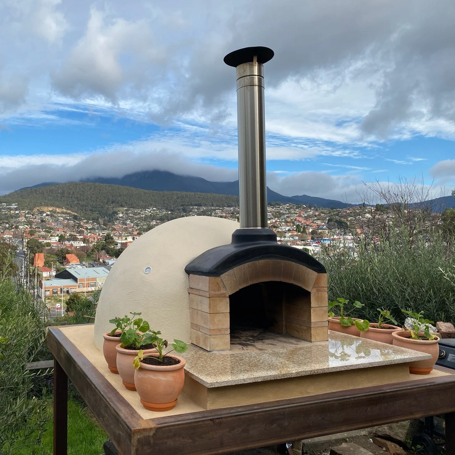 An outdoor pizza oven with a chimney on a stone countertop, surrounded by potted plants, overlooking a town and mountains under a partly cloudy sky.