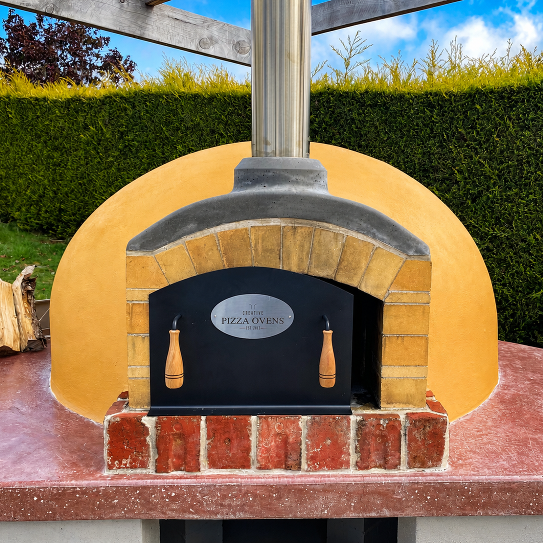A backyard pizza oven with a brick arch, black door, and a yellow stucco exterior, with a metal chimney on top, situated on a concrete counter, surrounded by green bushes and a blue sky.