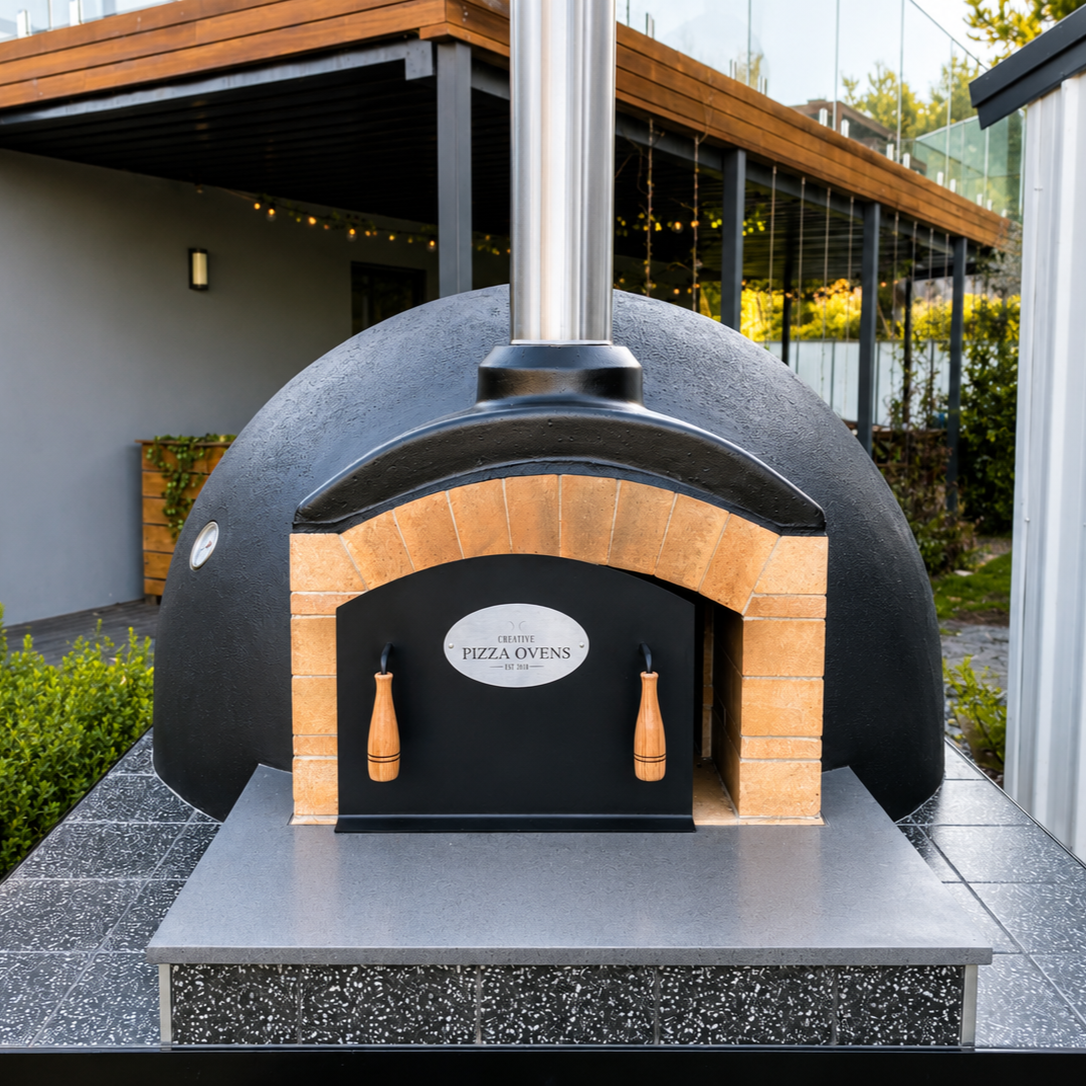 Outdoor pizza oven with a black dome, brick arch, and metal chimney, set on a tiled surface in a backyard.