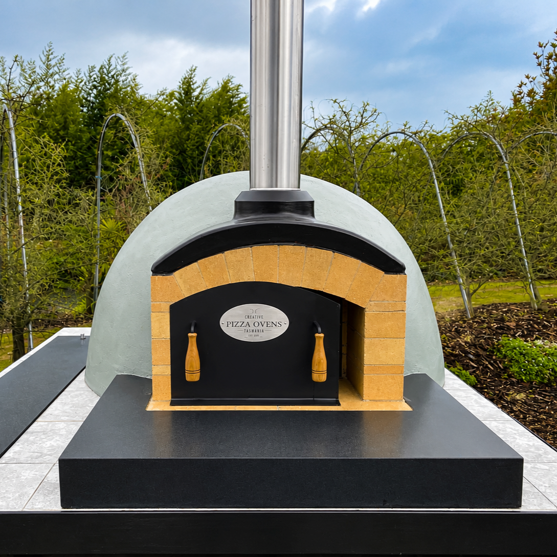 A pizza oven outdoors with a dome shape, brick arch entrance, and a metal chimney on top, surrounded by garden plants and trees.