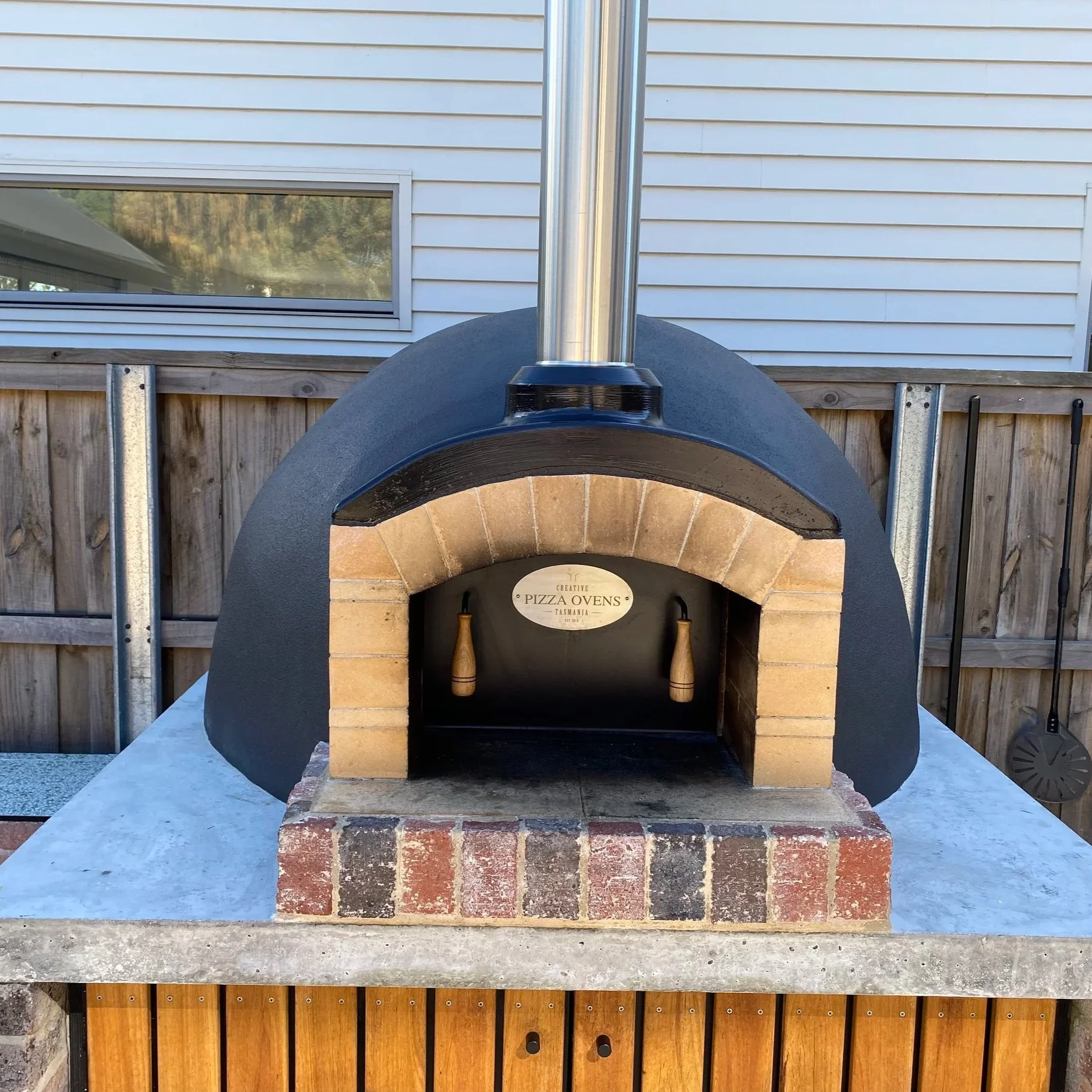An outdoor pizza oven with a brick interior and a black exterior, placed on a concrete and brick base, with a metal chimney on top, and wooden and metal tools beside it. A sign inside reads 'Creative Pizza Ovens Tasmania'.