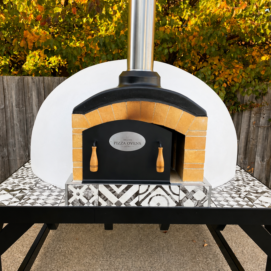 Outdoor pizza oven with a black door and a chimney, set on a patterned black, white, and gray tiled table, surrounded by a wooden fence and green foliage.