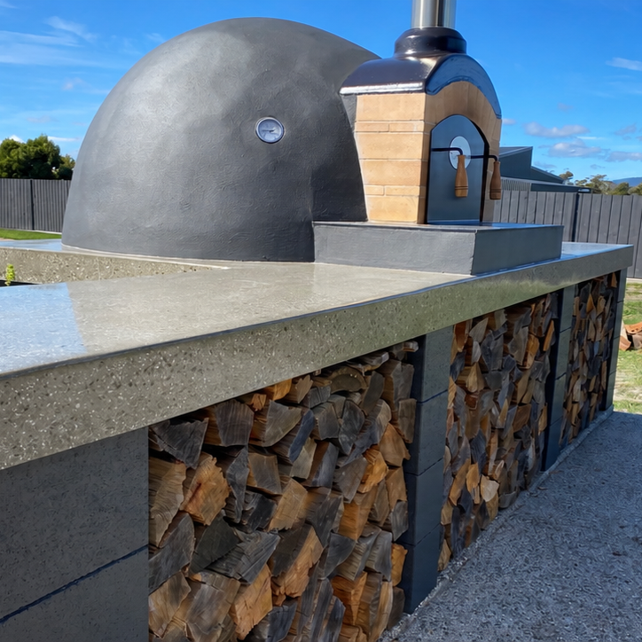 Outdoor stone pizza oven with a wood fire chamber, built on a countertop with a wood storage area underneath, and a clear sky in the background.